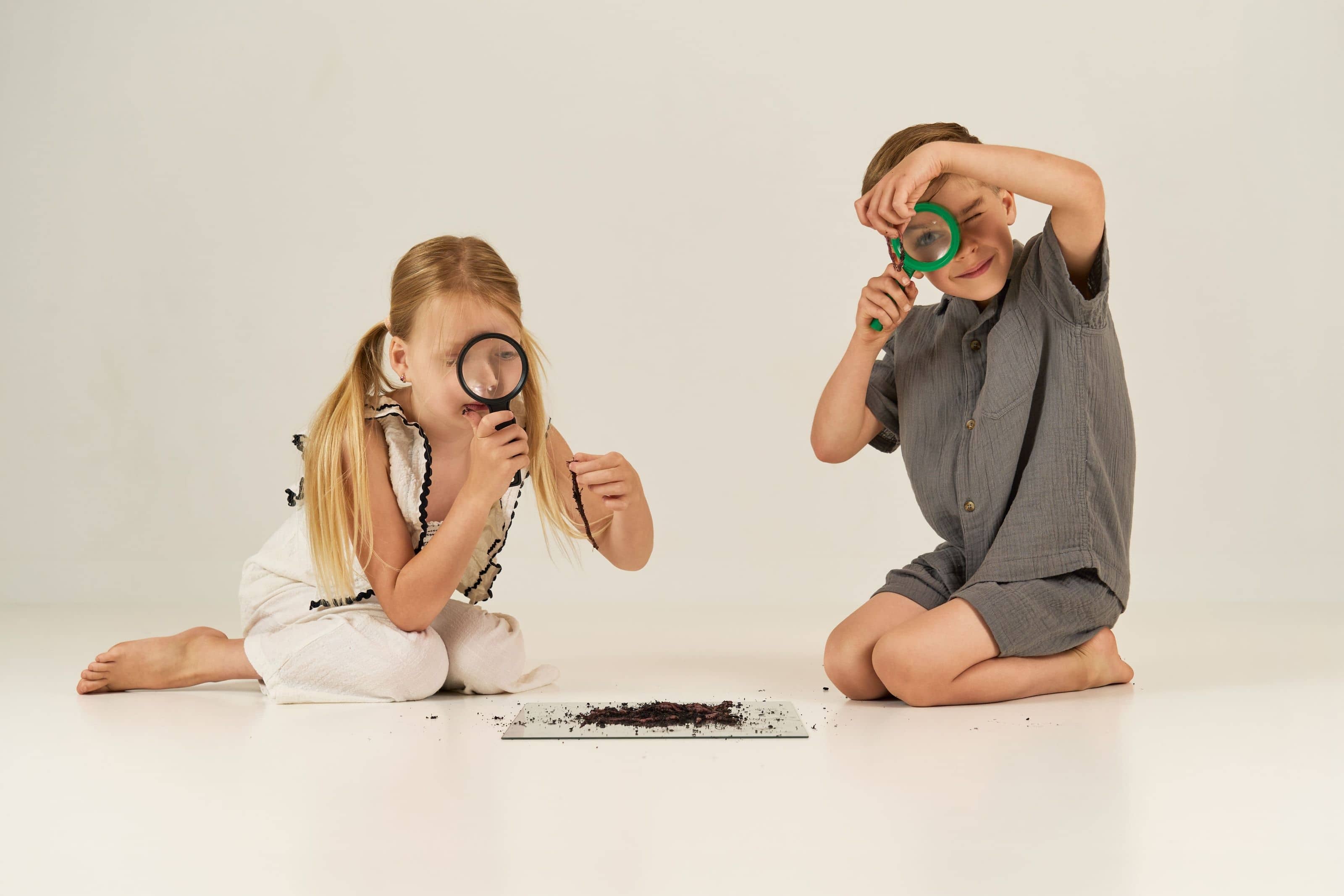 A girl and a boy sitting on the floor with magnifying glasses, examining a tray of soil between them