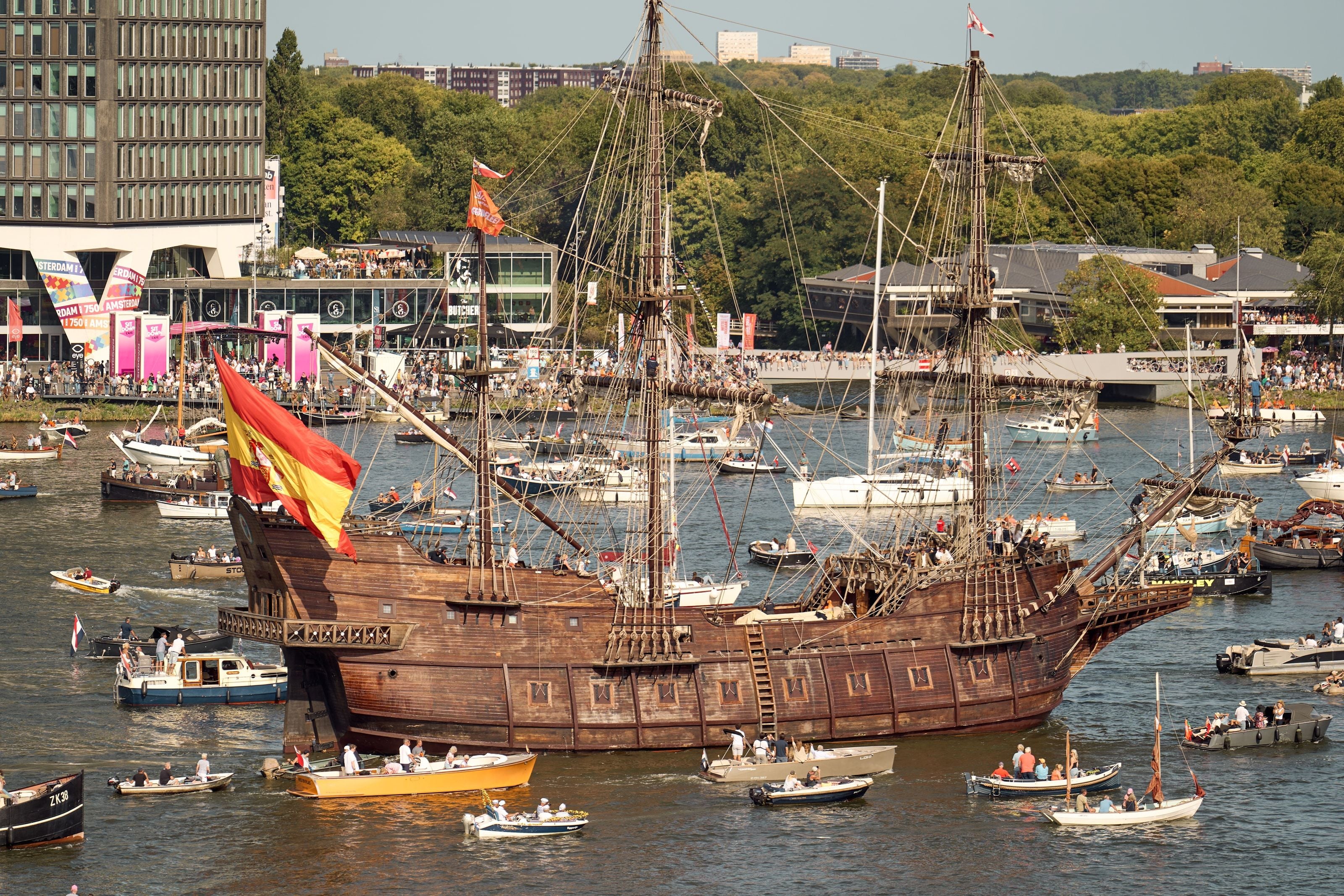 A wooden Spanish galleon flying a large Spanish flag on the IJ, small boats crowding around it and Amsterdam 750 banners on the quay behind