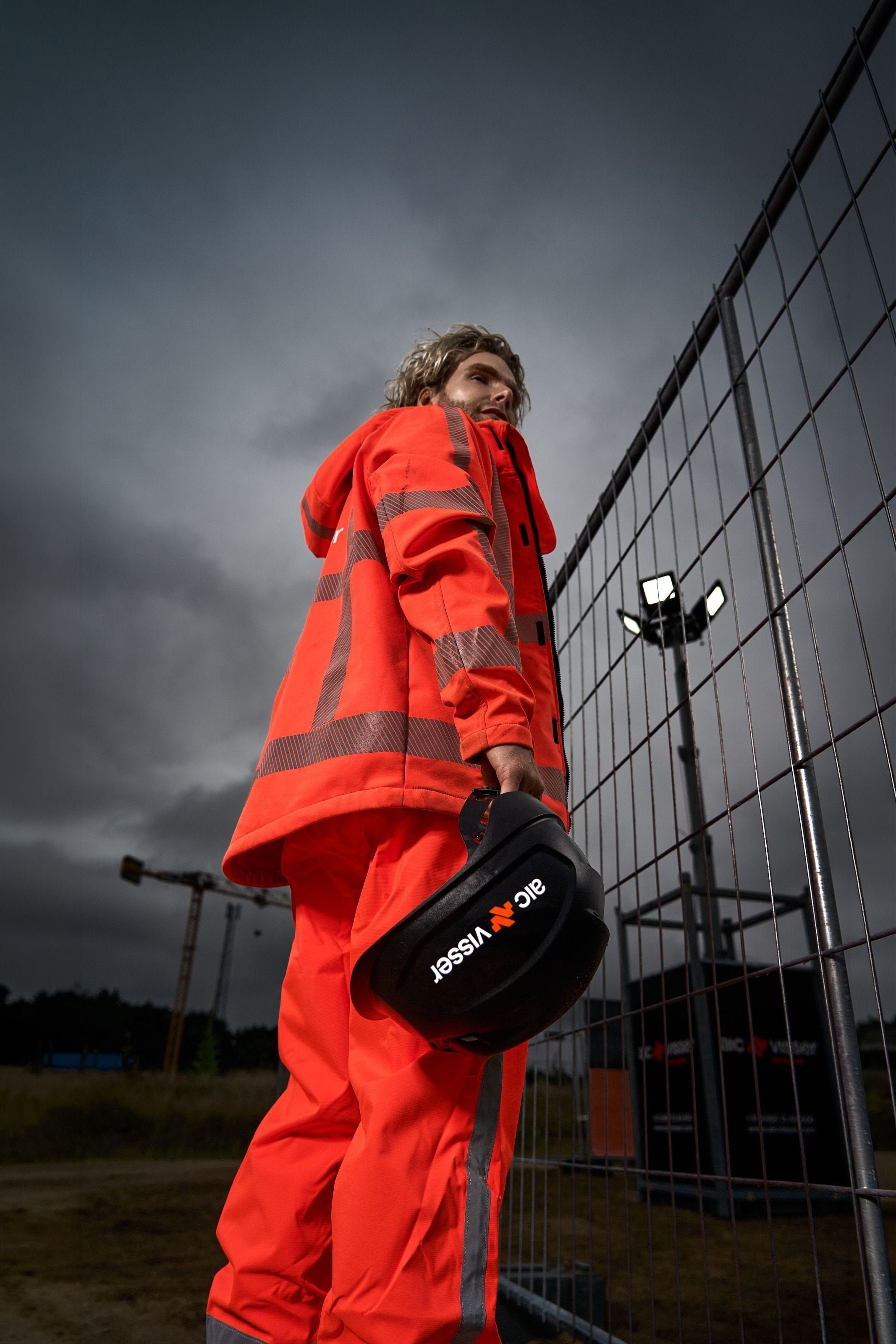 A worker in orange hi-vis seen from below, carrying an AIC Visser helmet past a site fence under a heavy sky