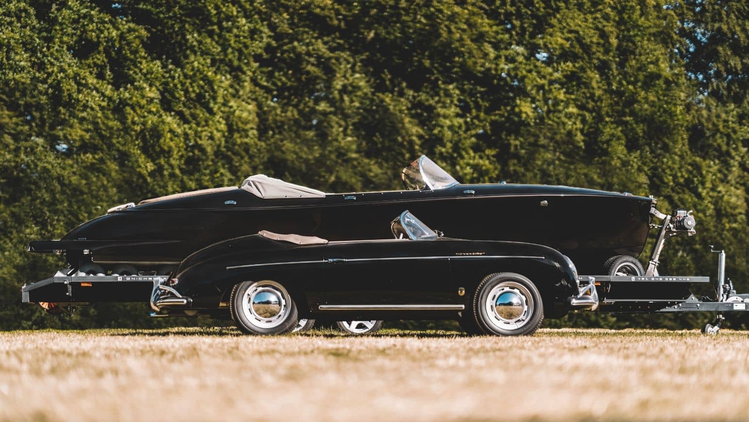 A black Porsche 356 Speedster parked in front of the Hermes Speedster on its trailer, on grass at a concours