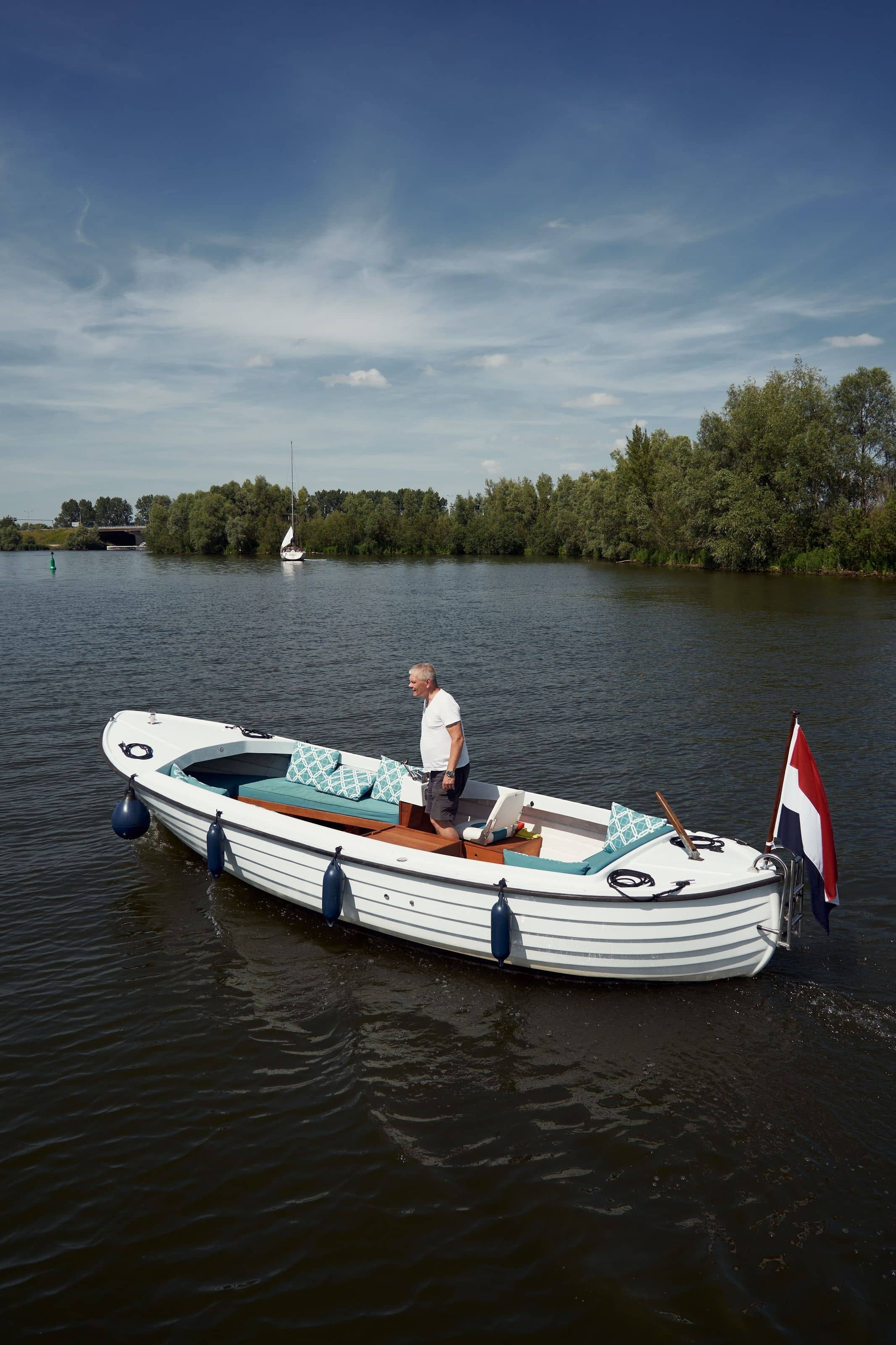 A white classic open sloep on the water near Harderwijk, its skipper standing amidships, a sailing boat and treeline behind