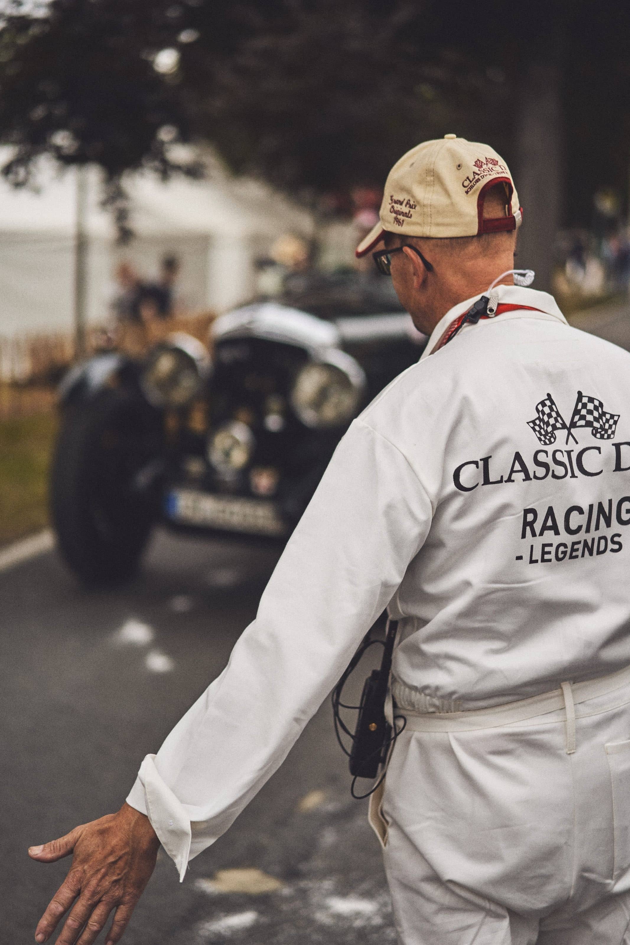 A marshal in a white "Classic Days Racing Legends" overall, radio on his belt, waving a vintage black car forward on the road