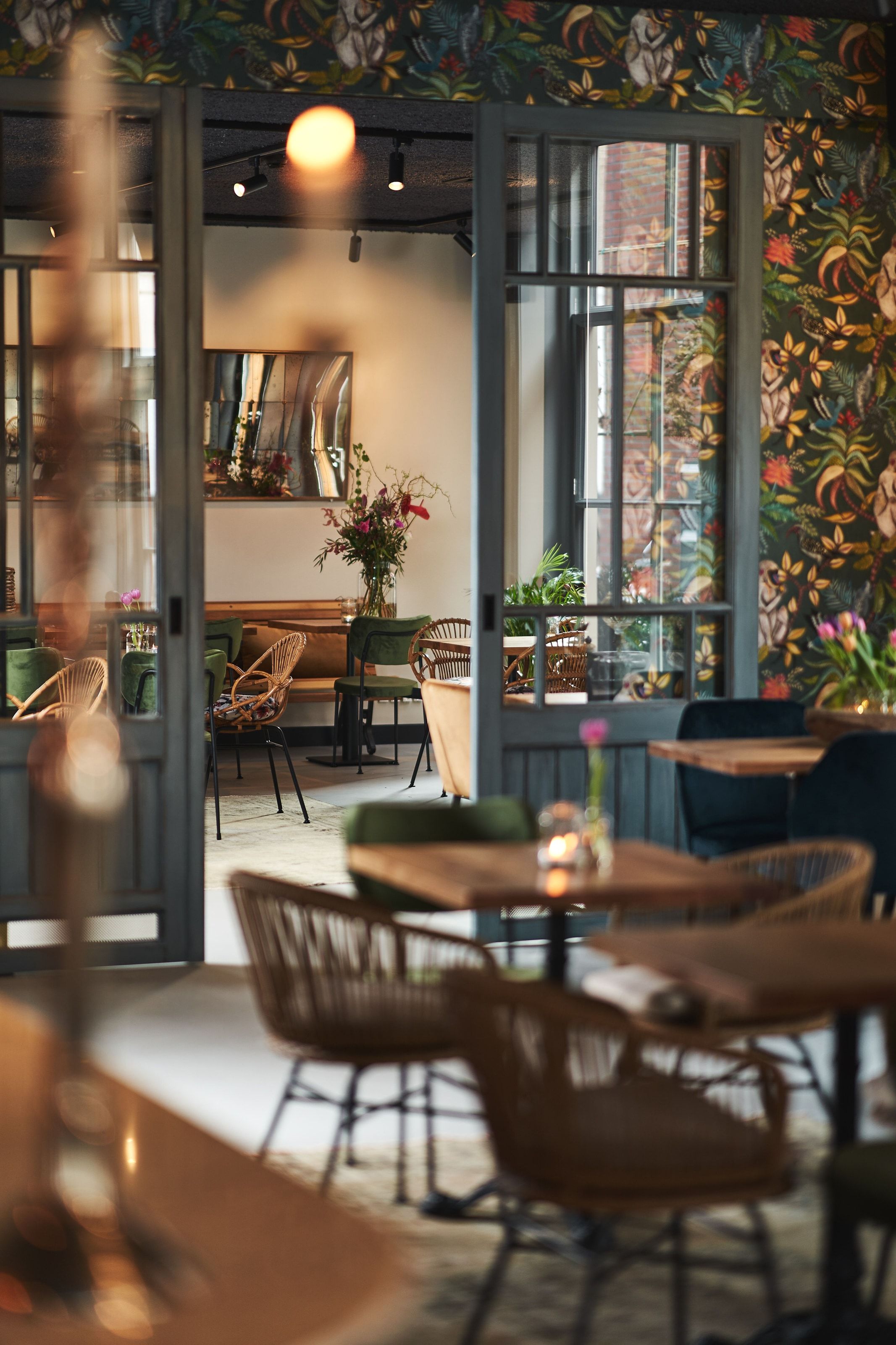Through open glazed doors into a second room, jungle-print wallpaper on one side and rattan and green velvet chairs beyond