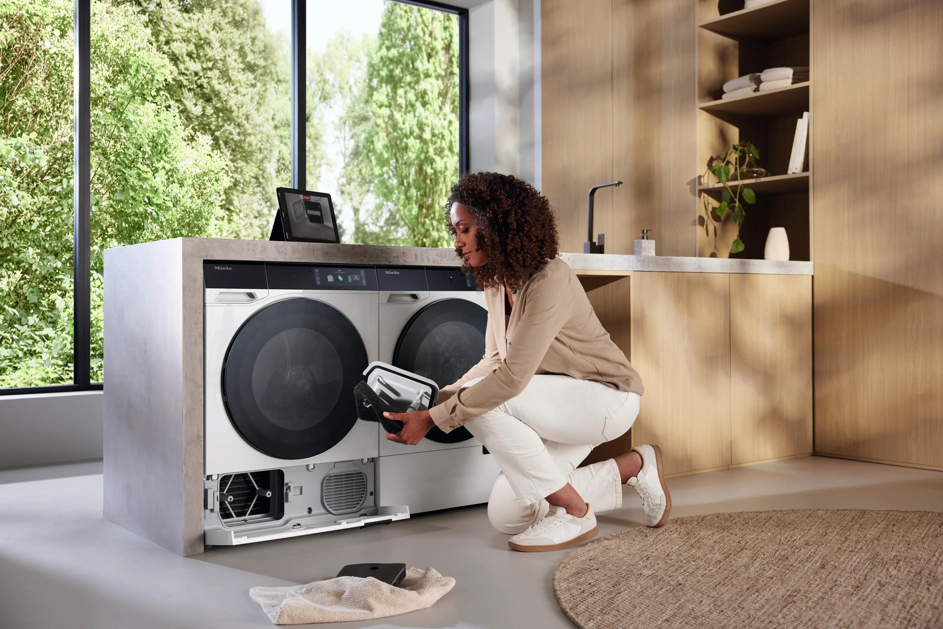A woman kneeling to remove the drain filter from a Miele washing machine, an iPad showing the app propped on the counter beside a second machine