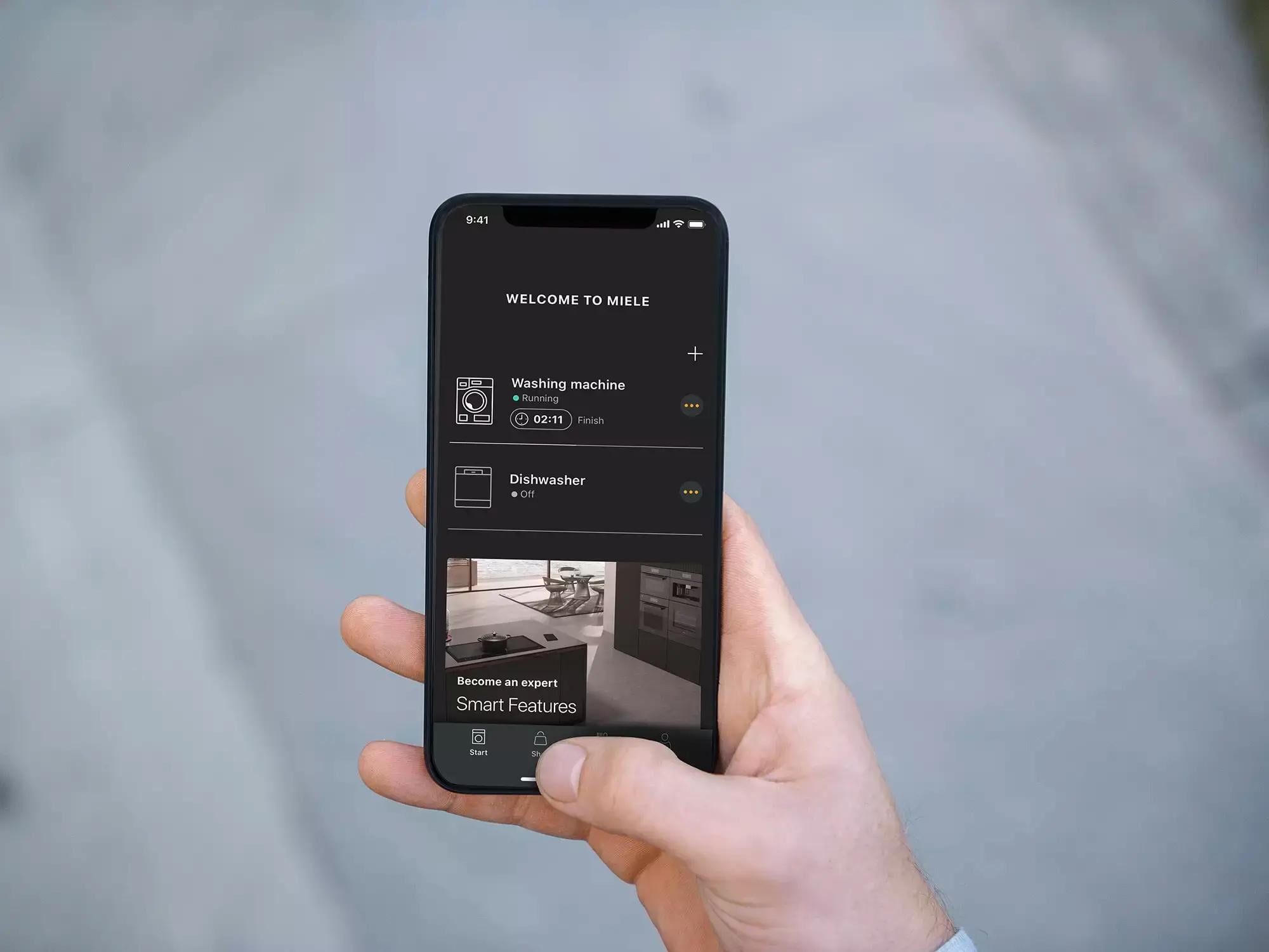 A hand holding a phone showing the Miele app’s home screen, a washing machine running and a dishwasher off