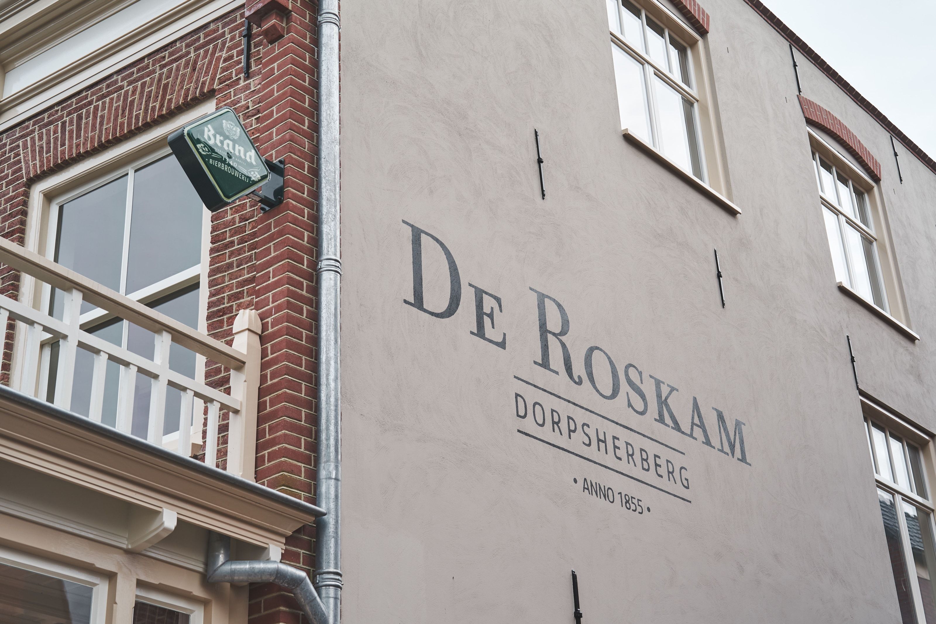 The painted gable end of De Roskam - the name, Dorpsherberg and Anno 1855 lettered on render, a Brand brewery sign on the corner