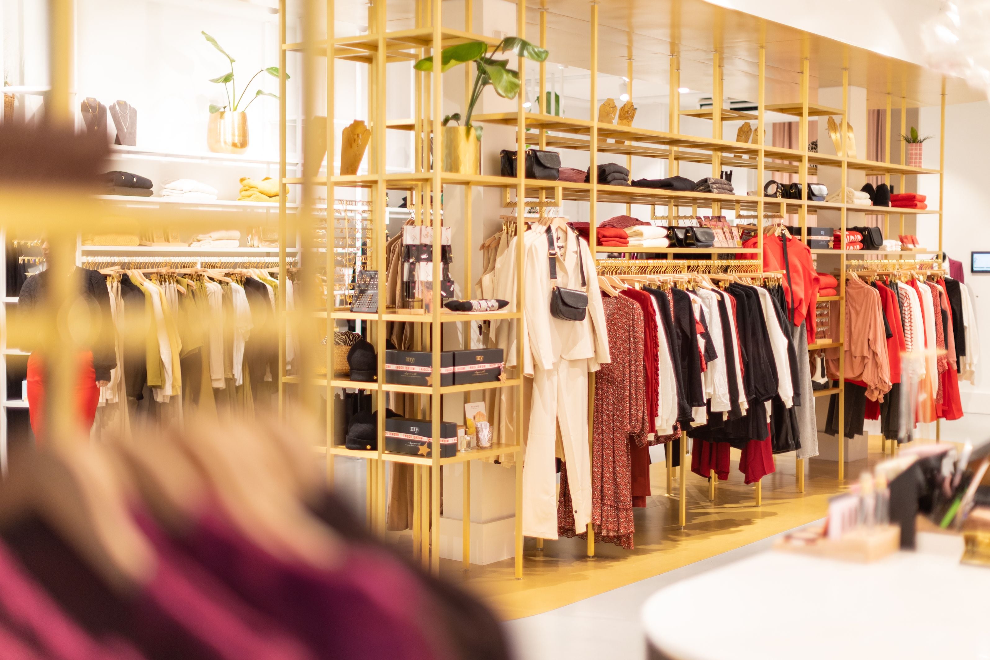 The interior of a My Jewellery boutique, gold shelving stacked with folded clothing, jewellery and handbags under a hanging plant