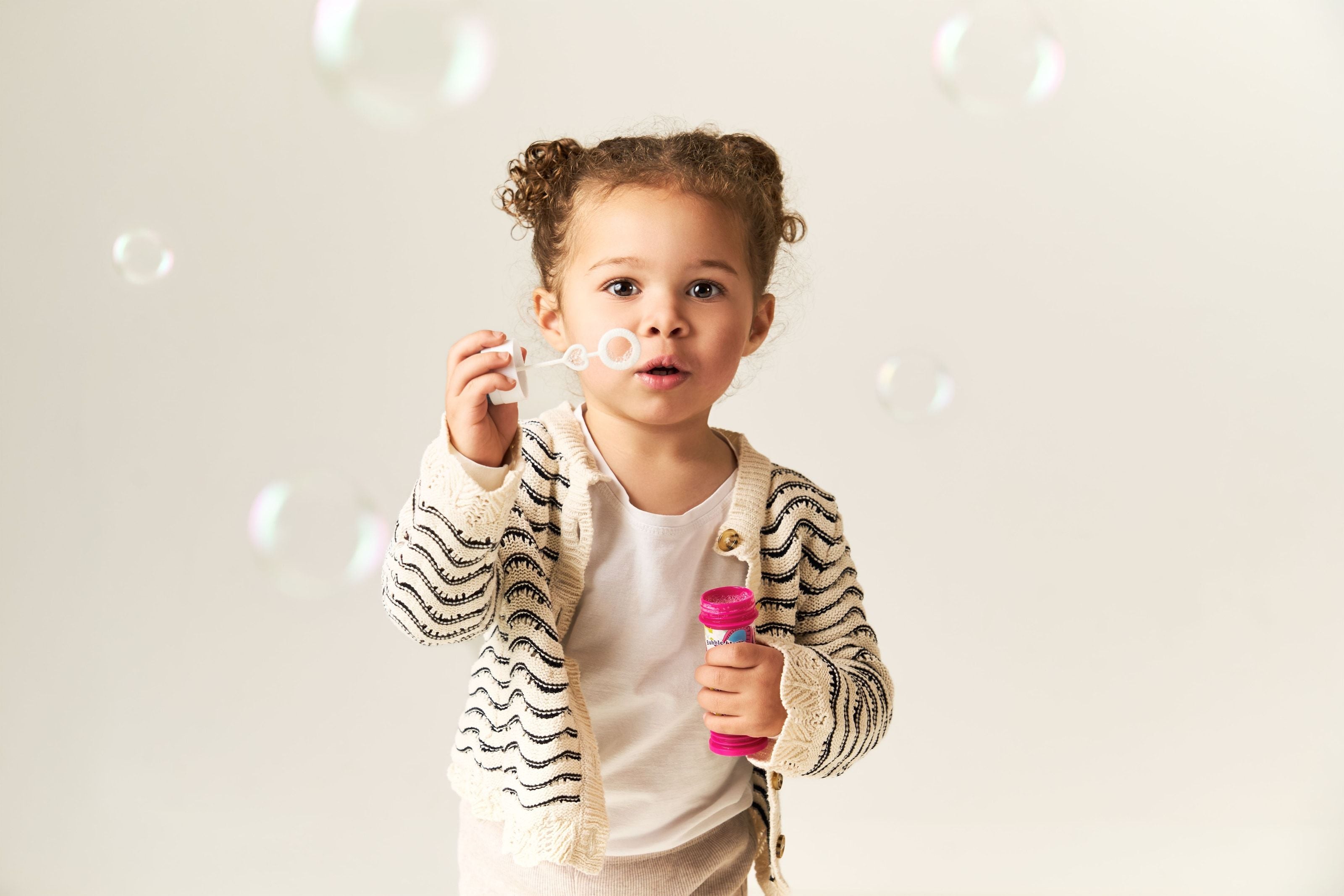A toddler in a striped cardigan blowing soap bubbles, bubbles drifting across the frame around her