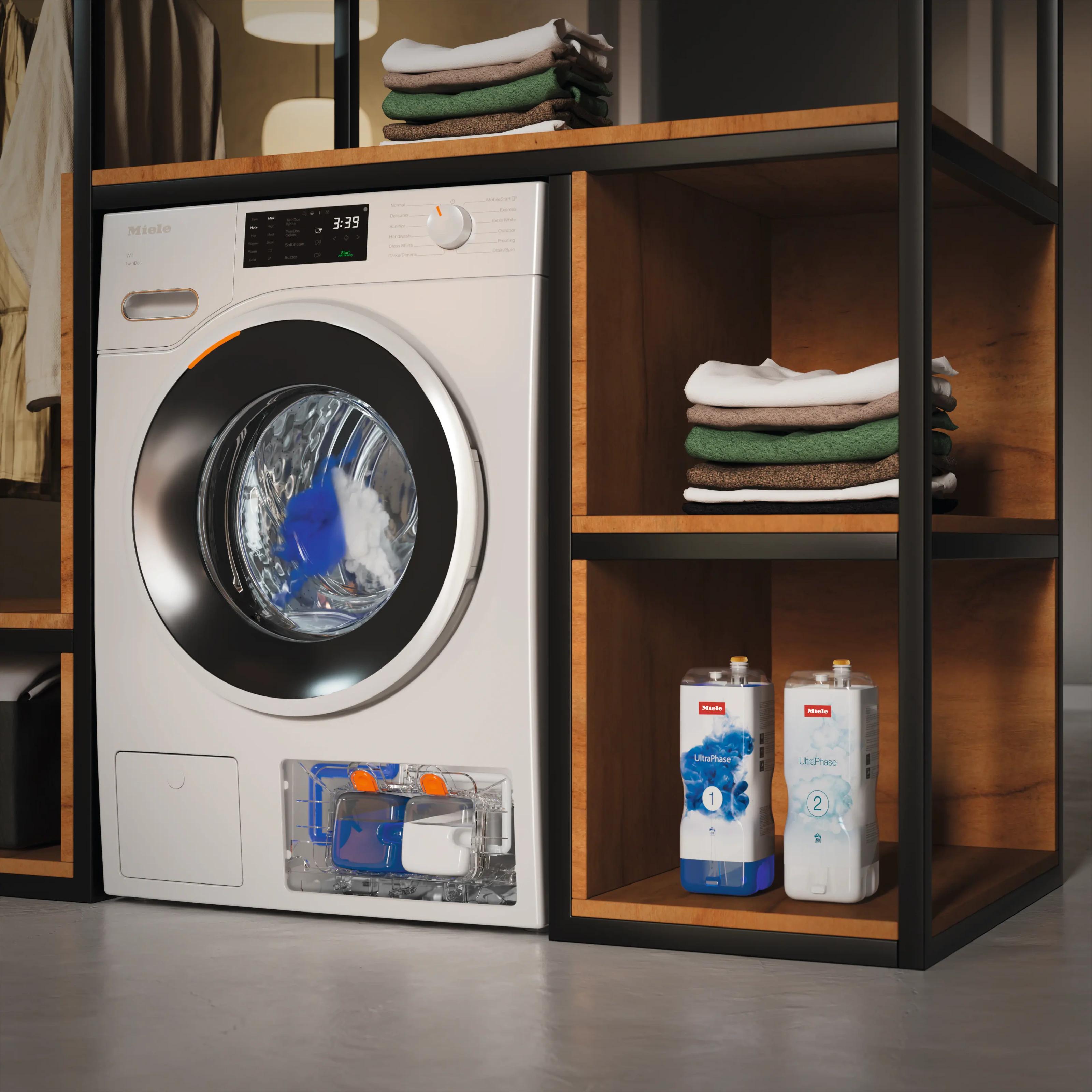 A Miele washing machine with its UltraPhase drawer open mid-cycle, two UltraPhase cartridges standing on a shelf beside folded laundry