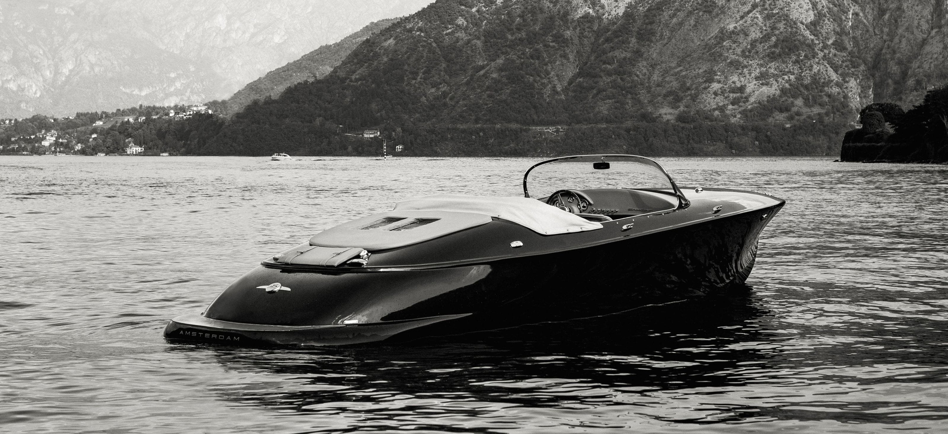 A dark Hermes Speedster at rest on Lake Como, steep wooded mountains behind it, in black and white