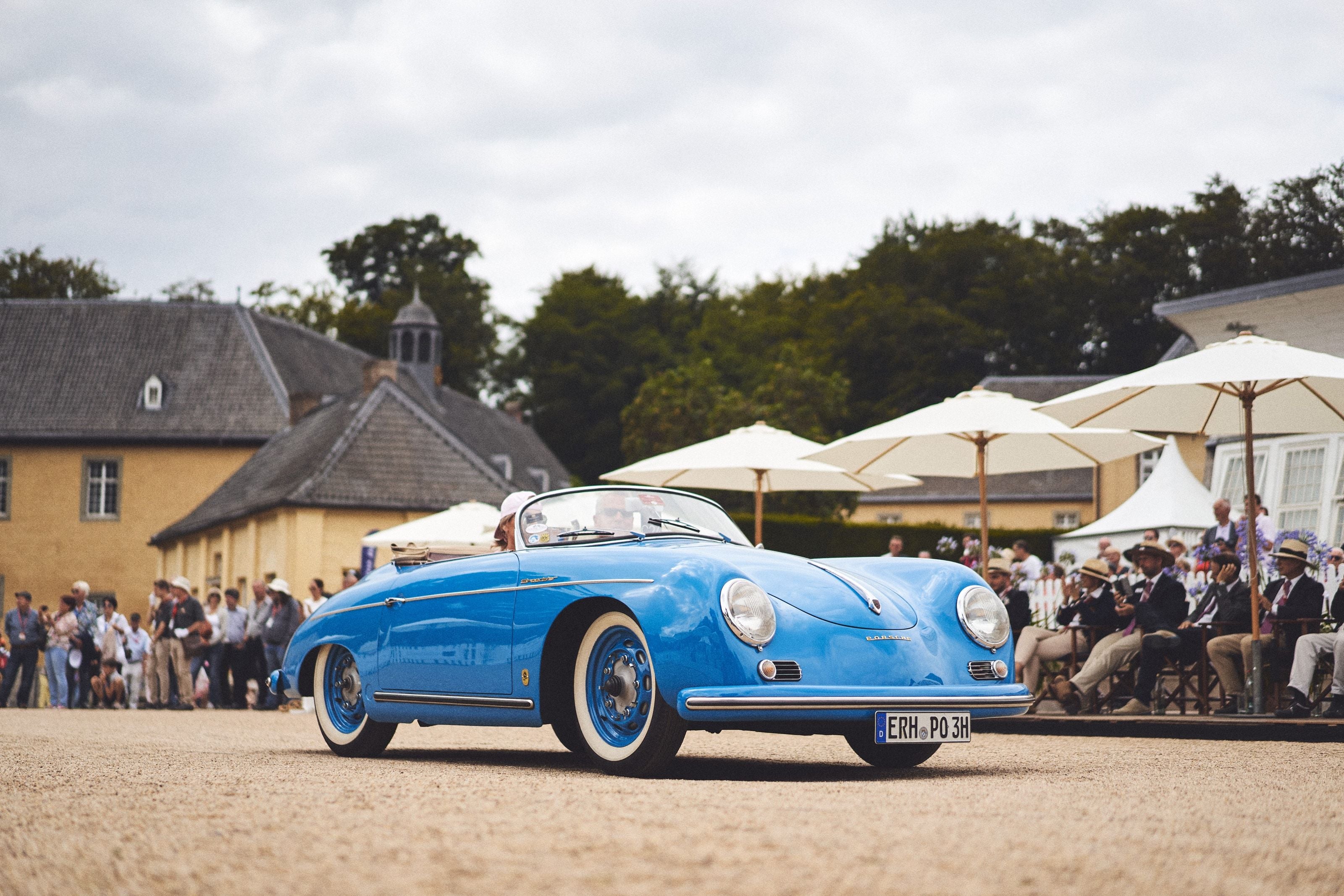 A pale blue Porsche 356 Speedster driving past a line of seated spectators, the buildings of Schloss Dyck and white umbrellas behind them