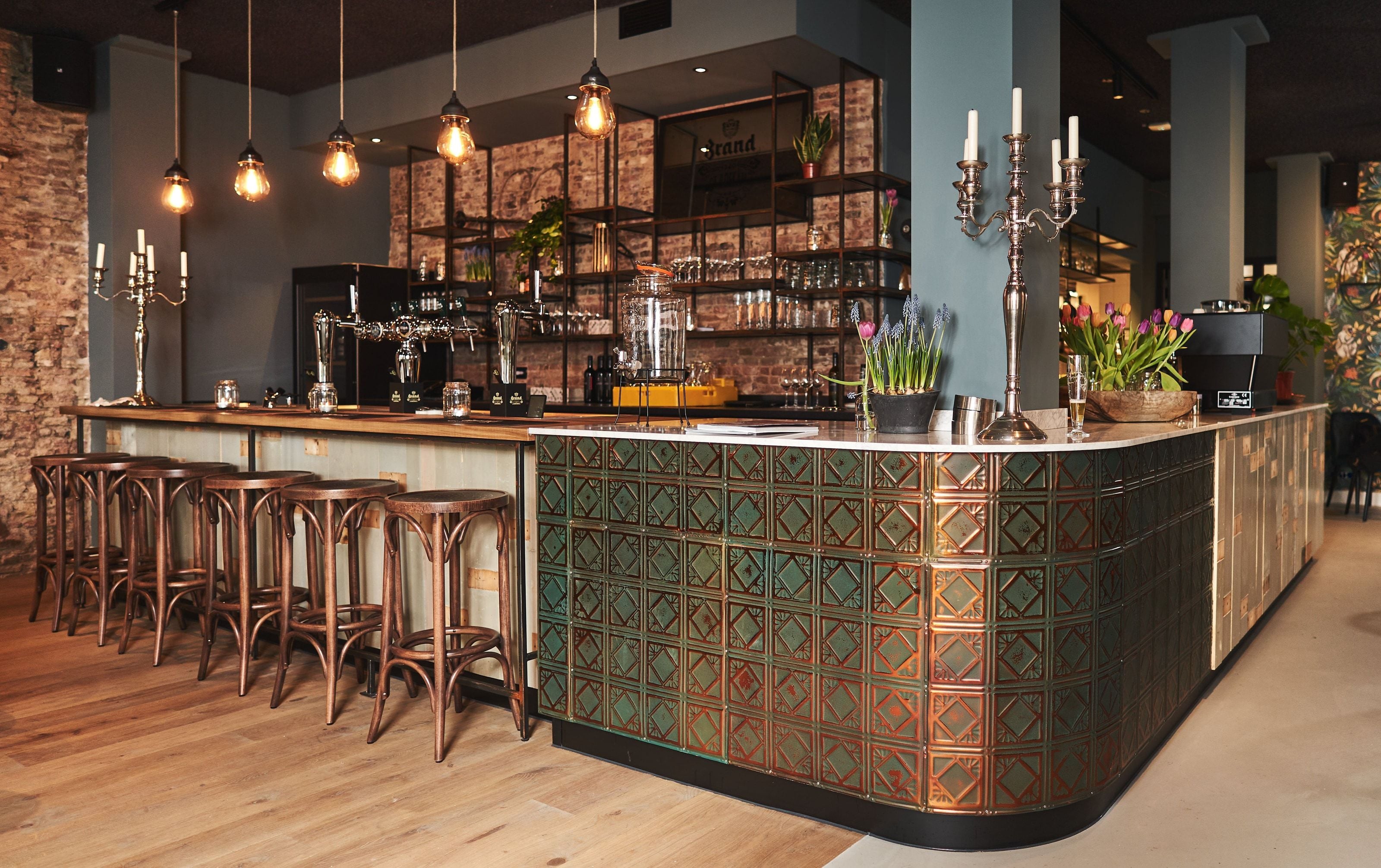 The bar seen down its length, filament bulbs hanging above it, a silver candelabra and pots of tulips on the tiled counter