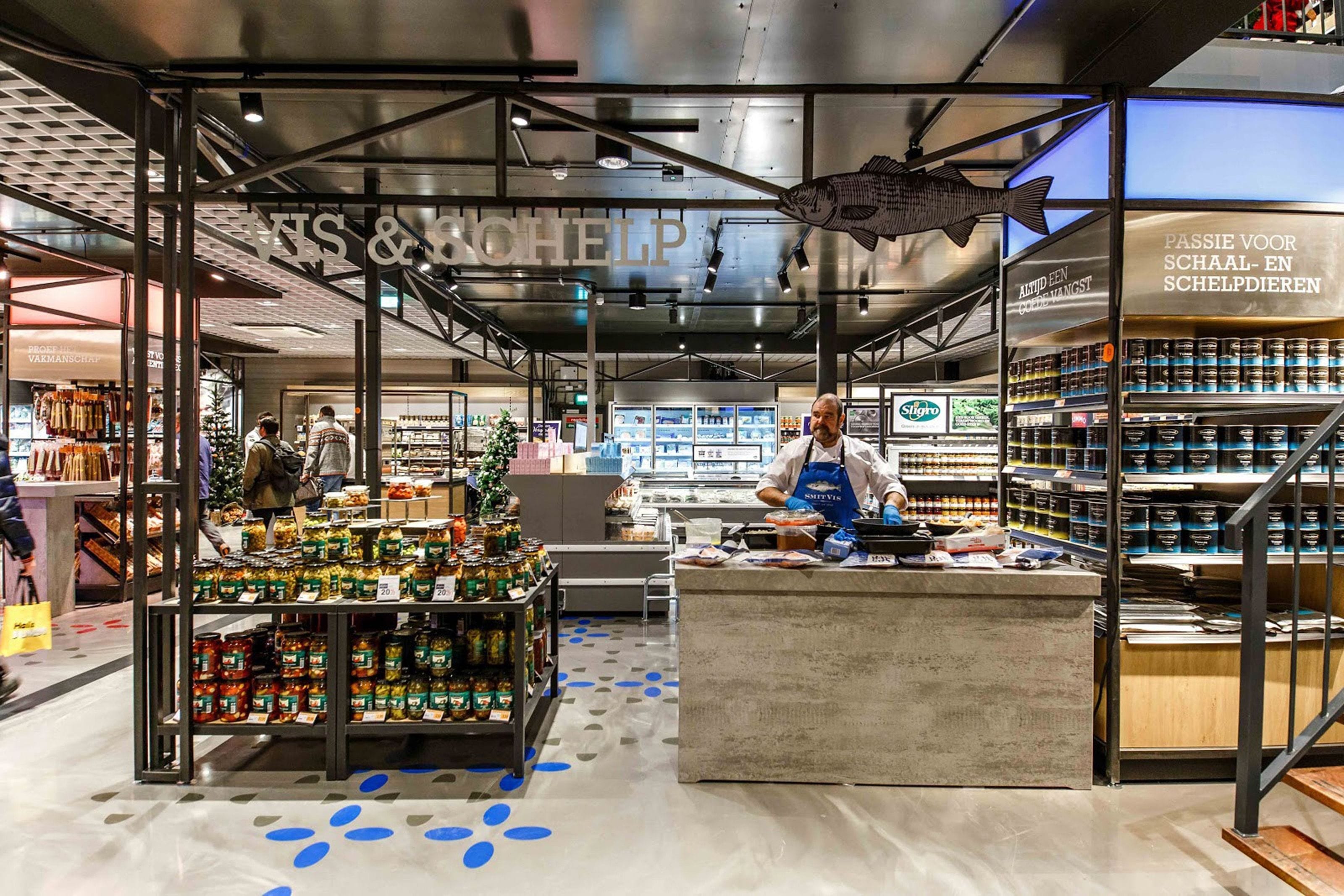 Inside a Sligro wholesale store, a fishmonger working the fresh fish counter with shelves of preserves behind him