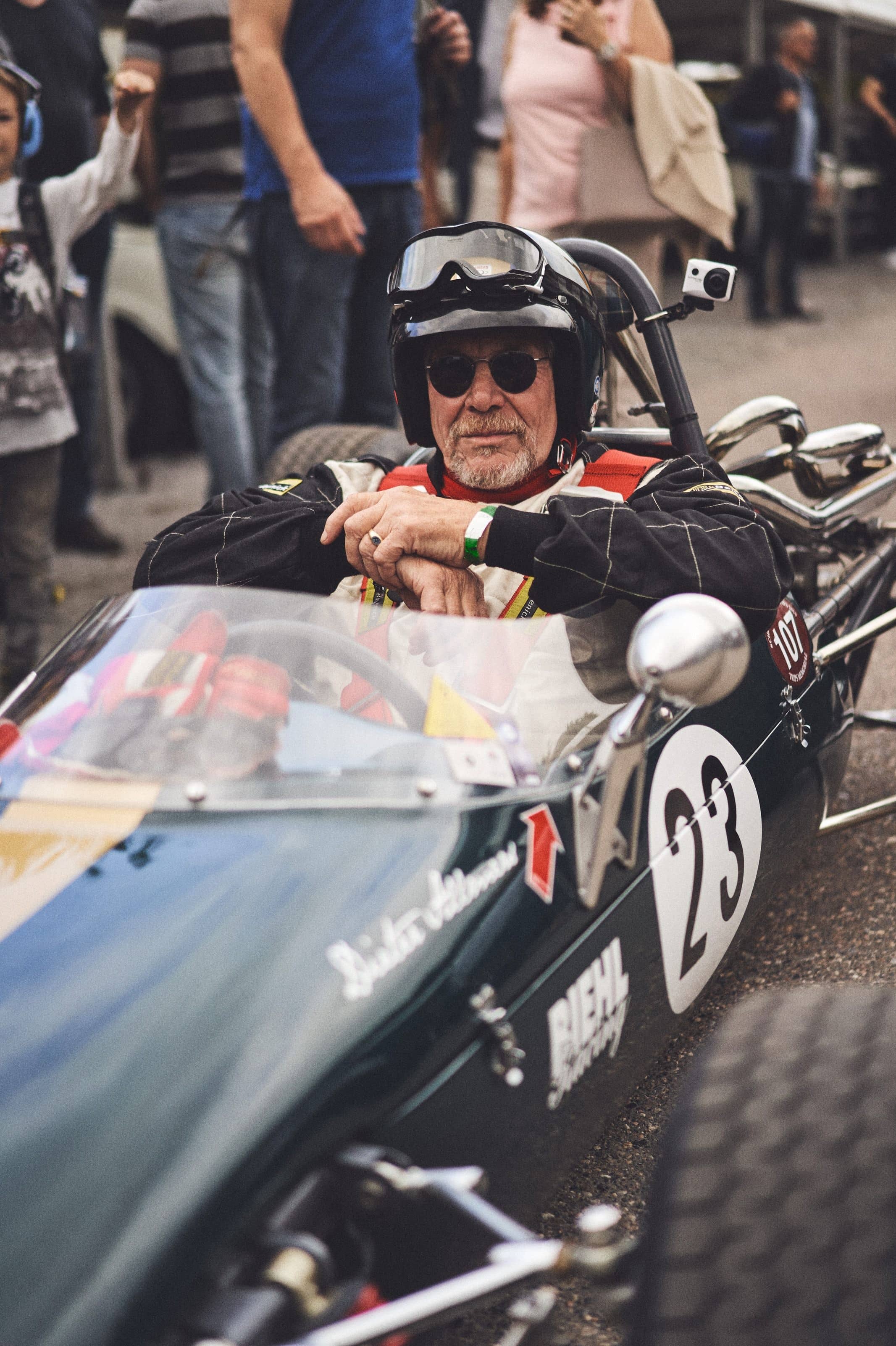 A driver in sunglasses and an open-face helmet, goggles pushed up, leaning on the cockpit edge of a dark green vintage single-seater