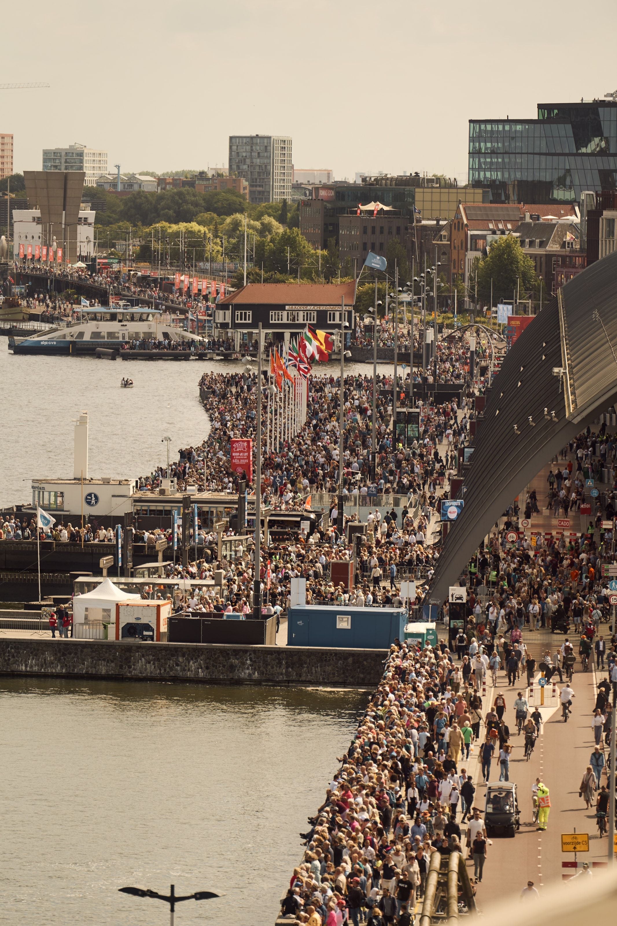 Crowds packed solid along the IJ waterfront and streaming across a bridge, a GVB ferry crossing behind them