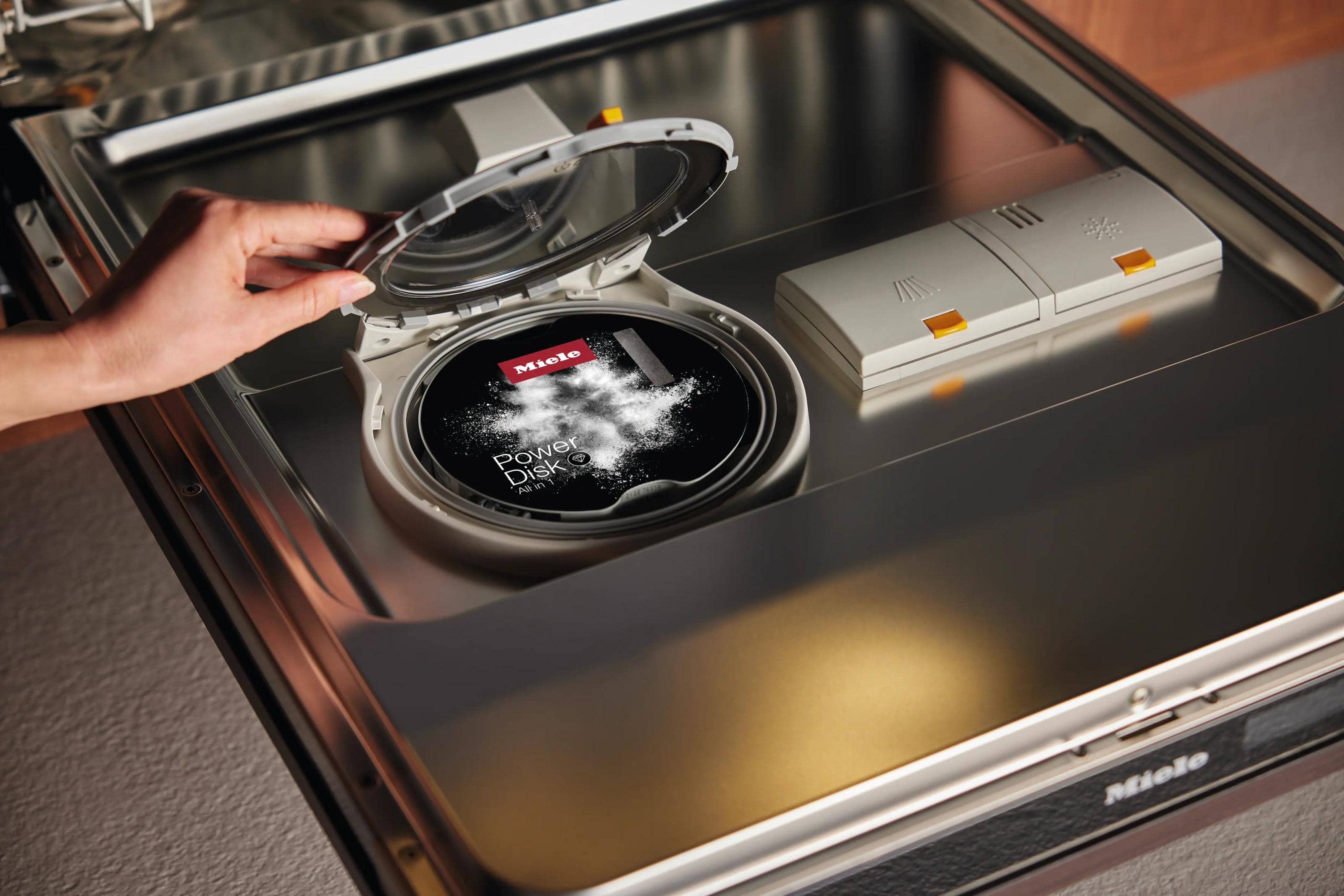 A hand opening a dishwasher’s PowerDisk compartment, the disk dusted with detergent powder
