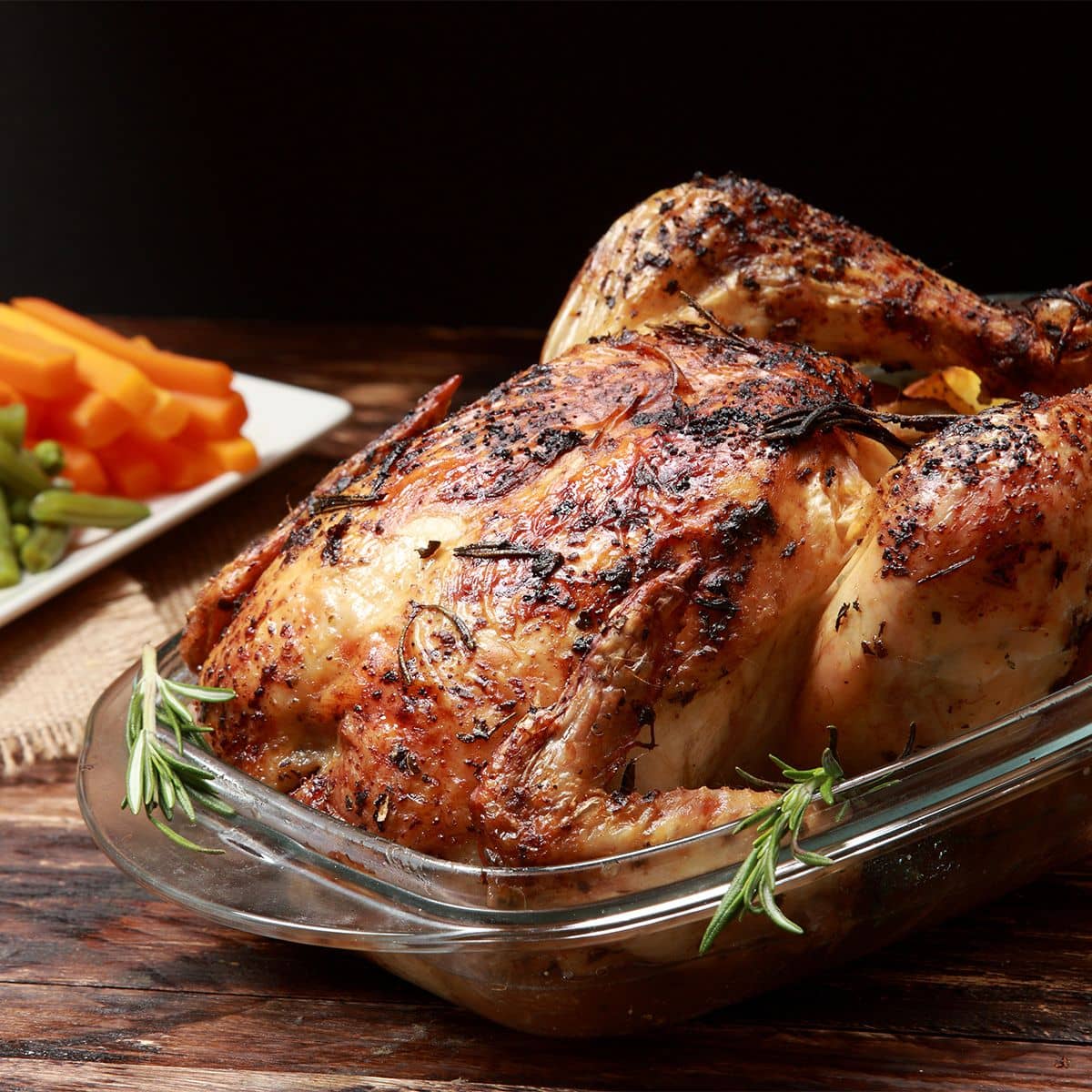 A whole roast chicken rubbed with herbs and rosemary sprigs, in a glass dish on a dark wood table, with carrot and celery sticks on a plate behind it