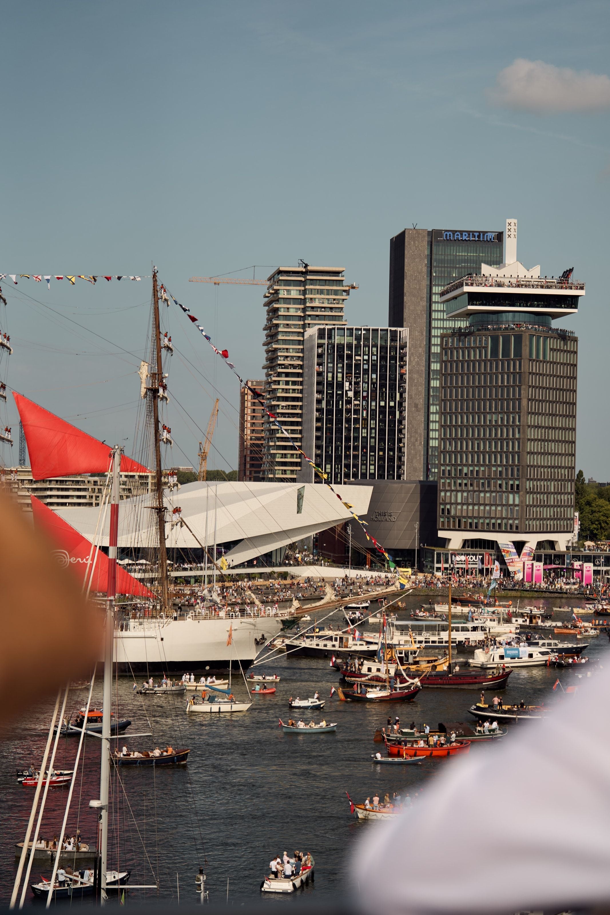 A tall ship passing the EYE film museum and the A’DAM Toren, its crew standing out along the yards in white, a red sail set forward