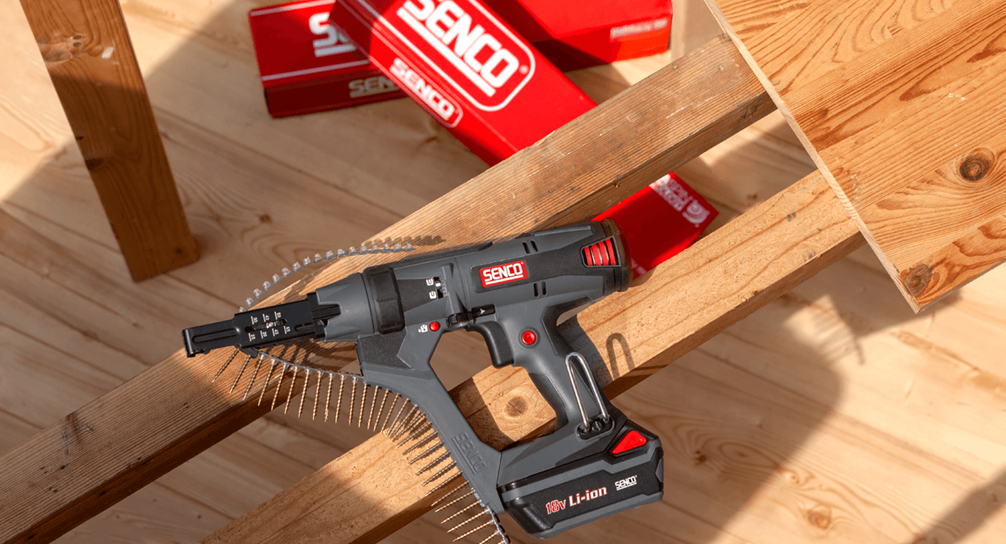 A grey and red Senco cordless fastening tool loaded with a coiled strip of screws, resting on timber decking with red Senco packaging boxes behind it