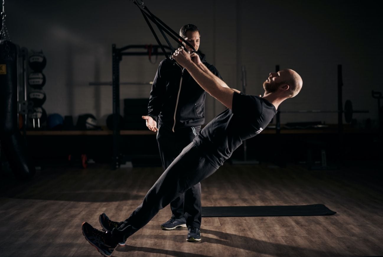 A client hanging back at full extension on suspension straps while a trainer spots him