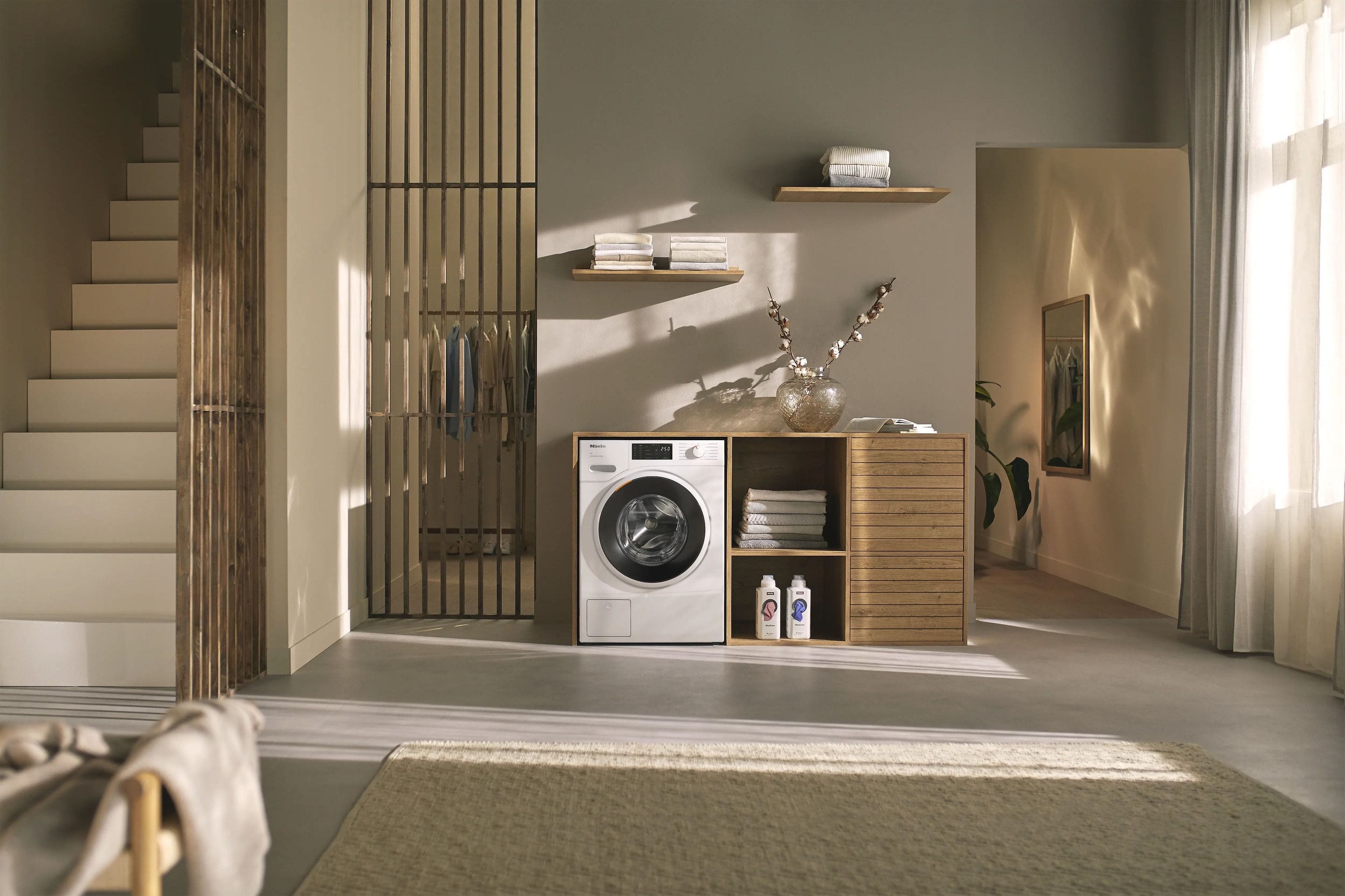 A Miele washing machine built into oak cabinetry in a sunlit hallway, laundry folded on the shelves beside it