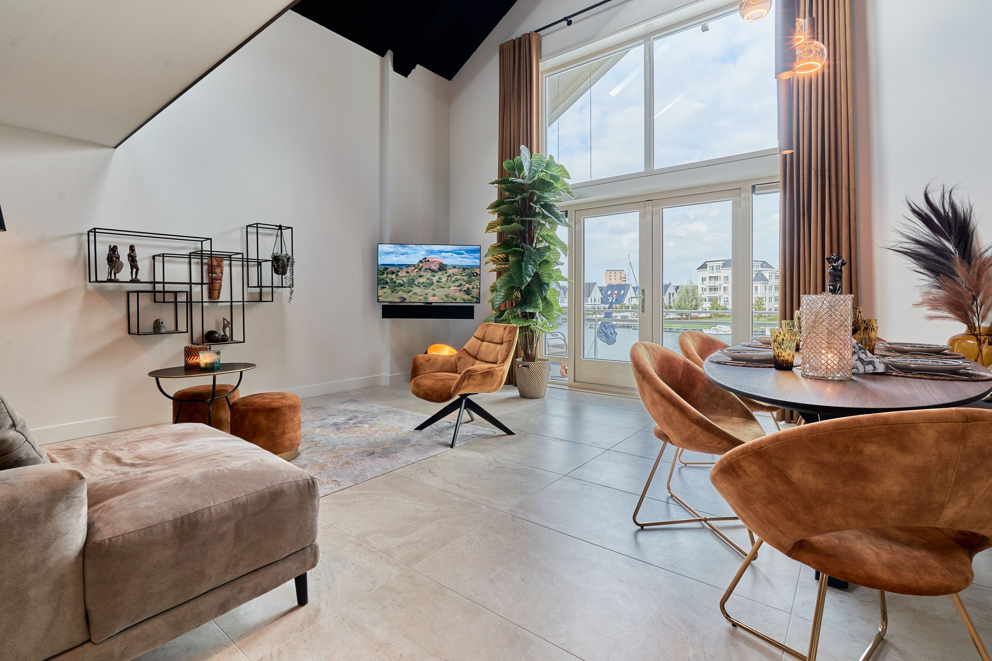 The living room of the Nomad Water Lodge under a pitched ceiling, rust velvet chairs at a laid dining table and floor-to-ceiling glass onto the marina
