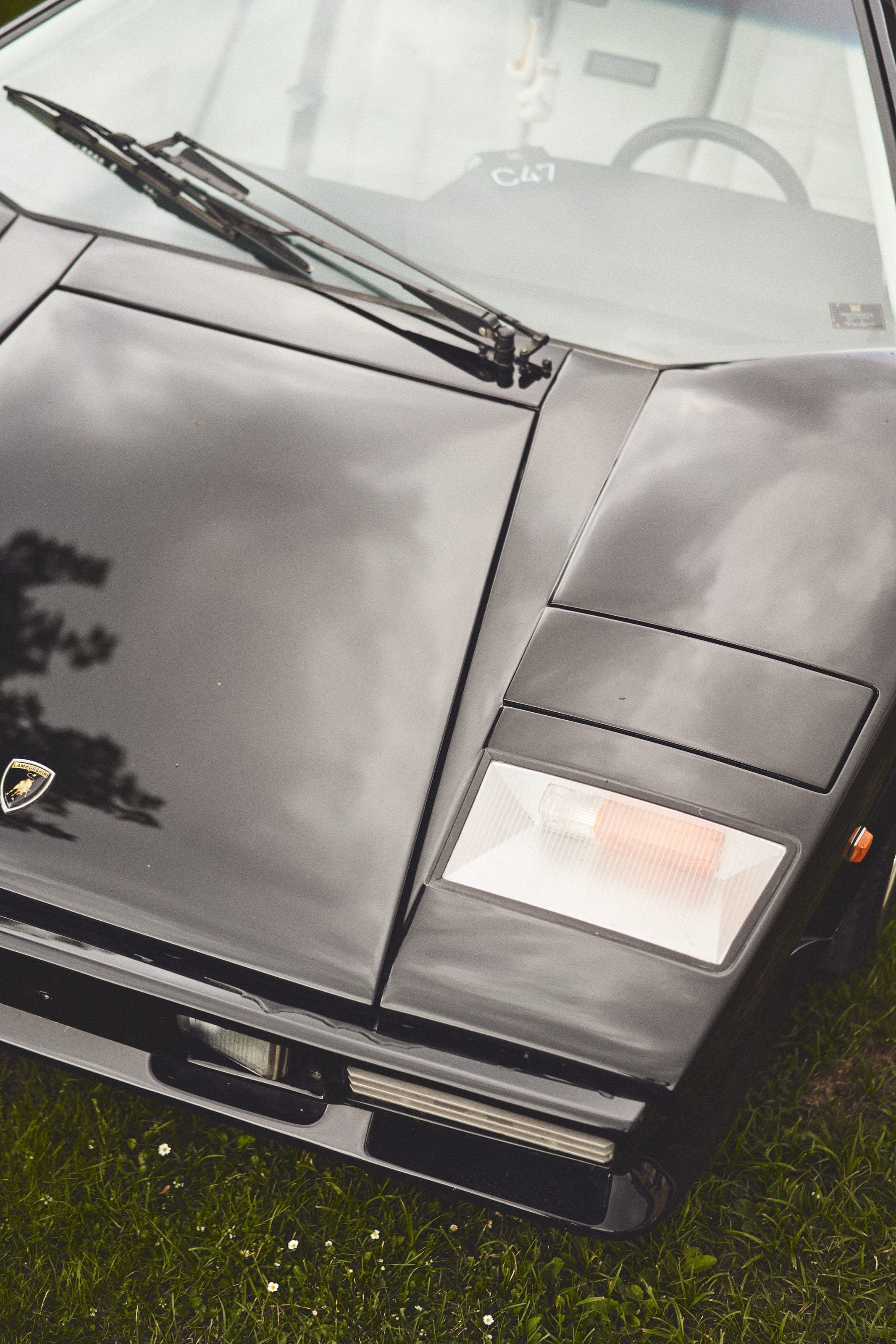 The low, wedge-shaped nose of a dark grey Lamborghini Countach, shot from above, its pop-up headlight raised, parked on grass