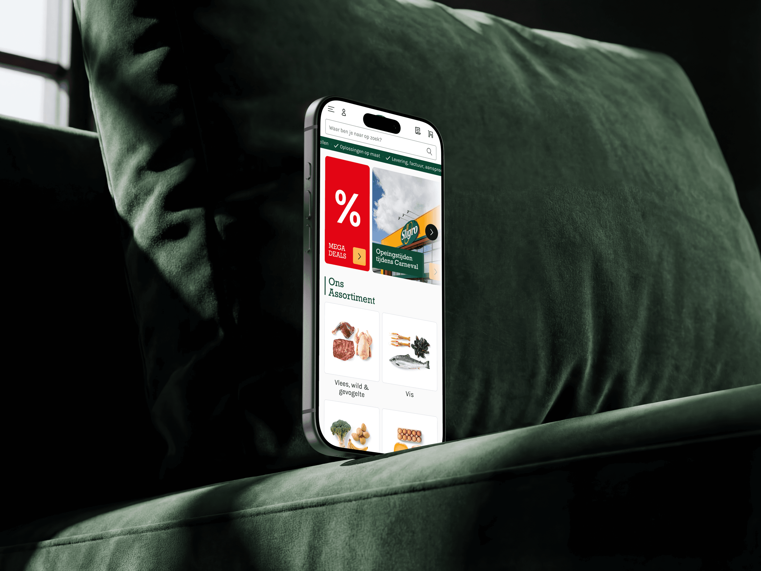 An iPhone upright on a dark green sofa showing the Sligro app home screen, a Mega Deals panel above the assortment categories