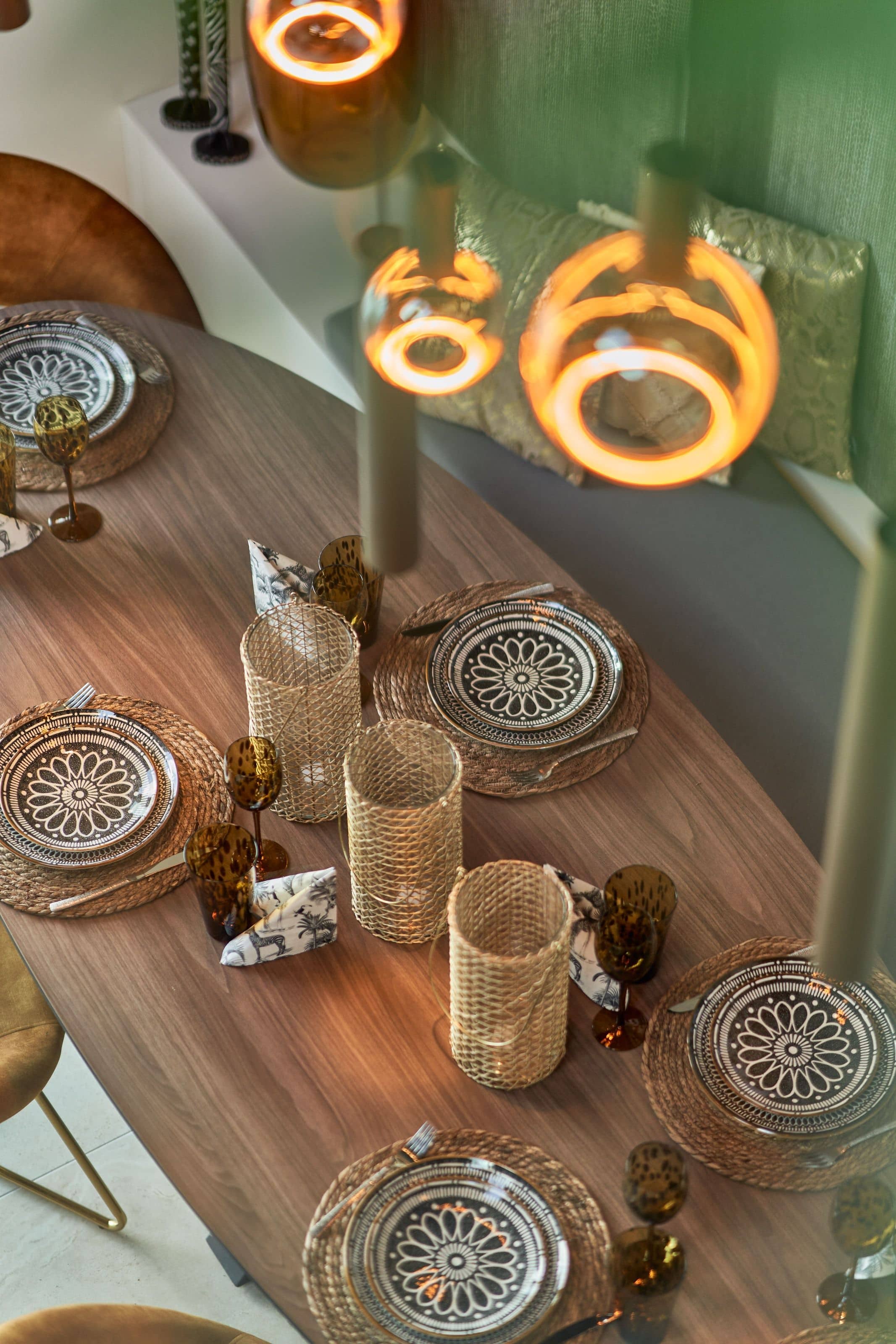 A laid table from above - patterned plates on rattan mats, amber tortoiseshell glasses, ring pendants glowing against a sage-green wall