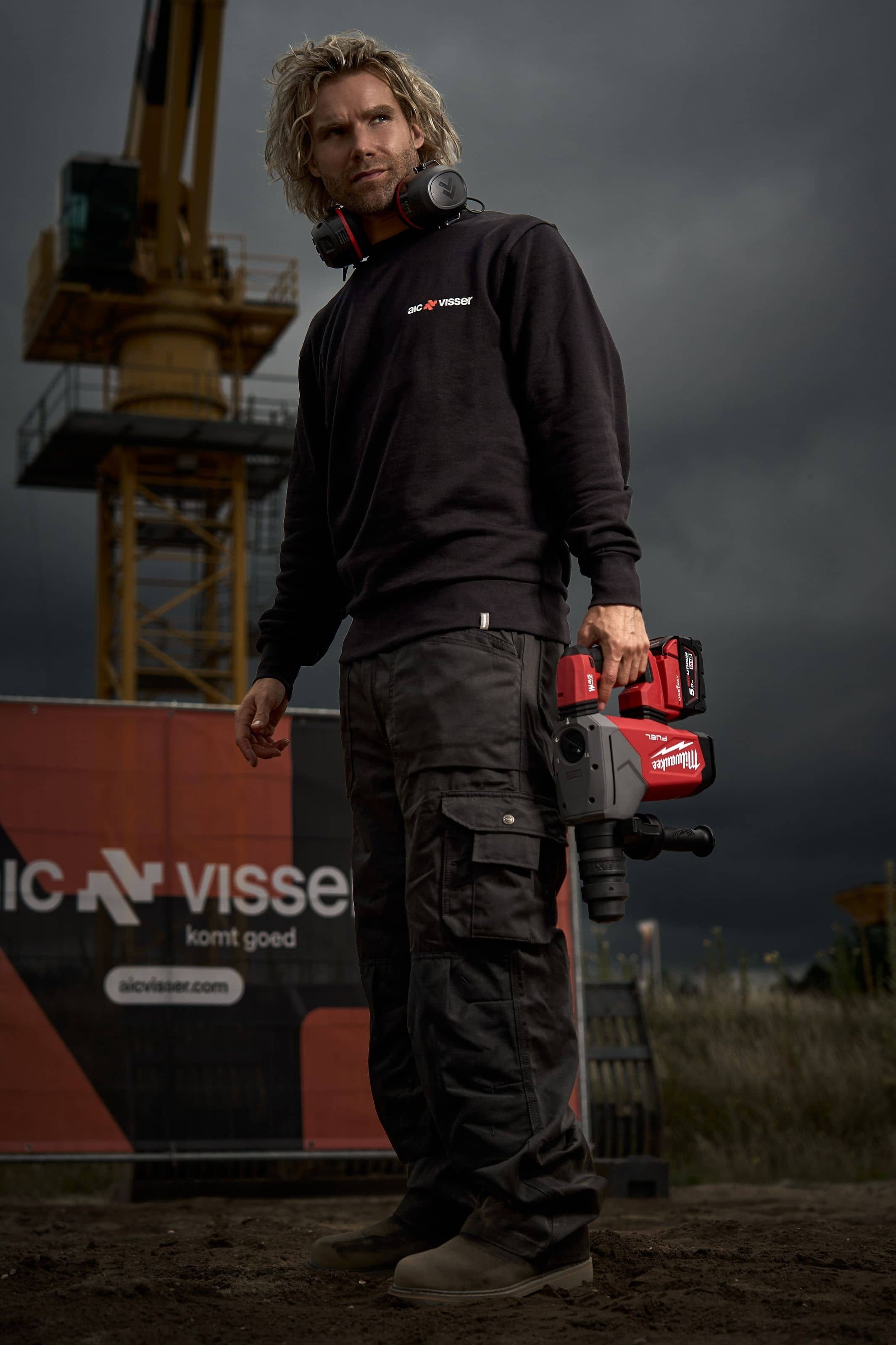 A man in an AIC Visser sweatshirt holding a breaker, a yellow crane and the company's hoarding behind him