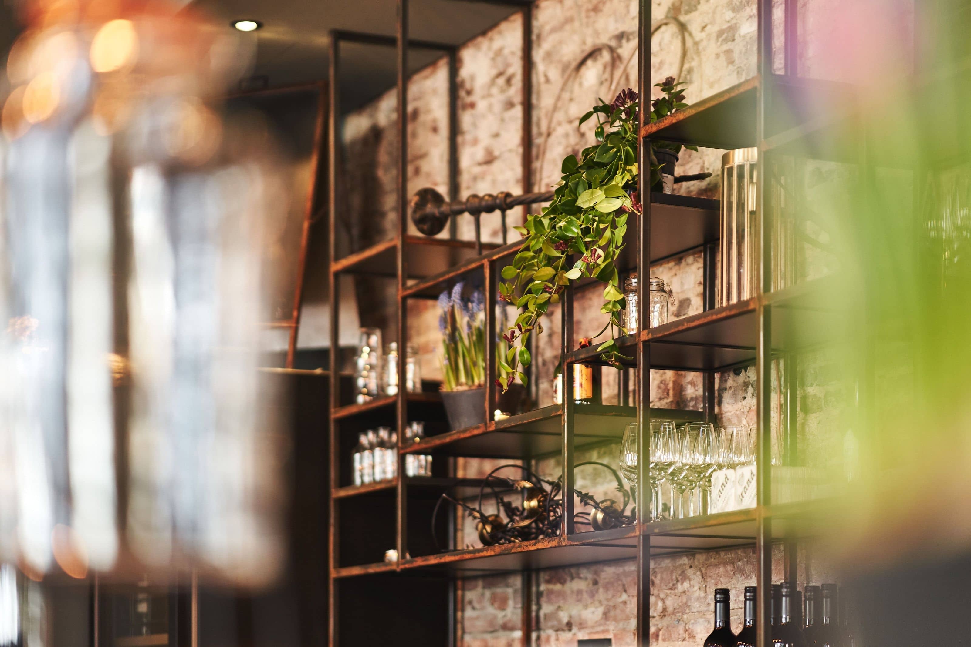 A steel shelf unit against bare brick behind the bar, trailing plants, wine glasses and bottles