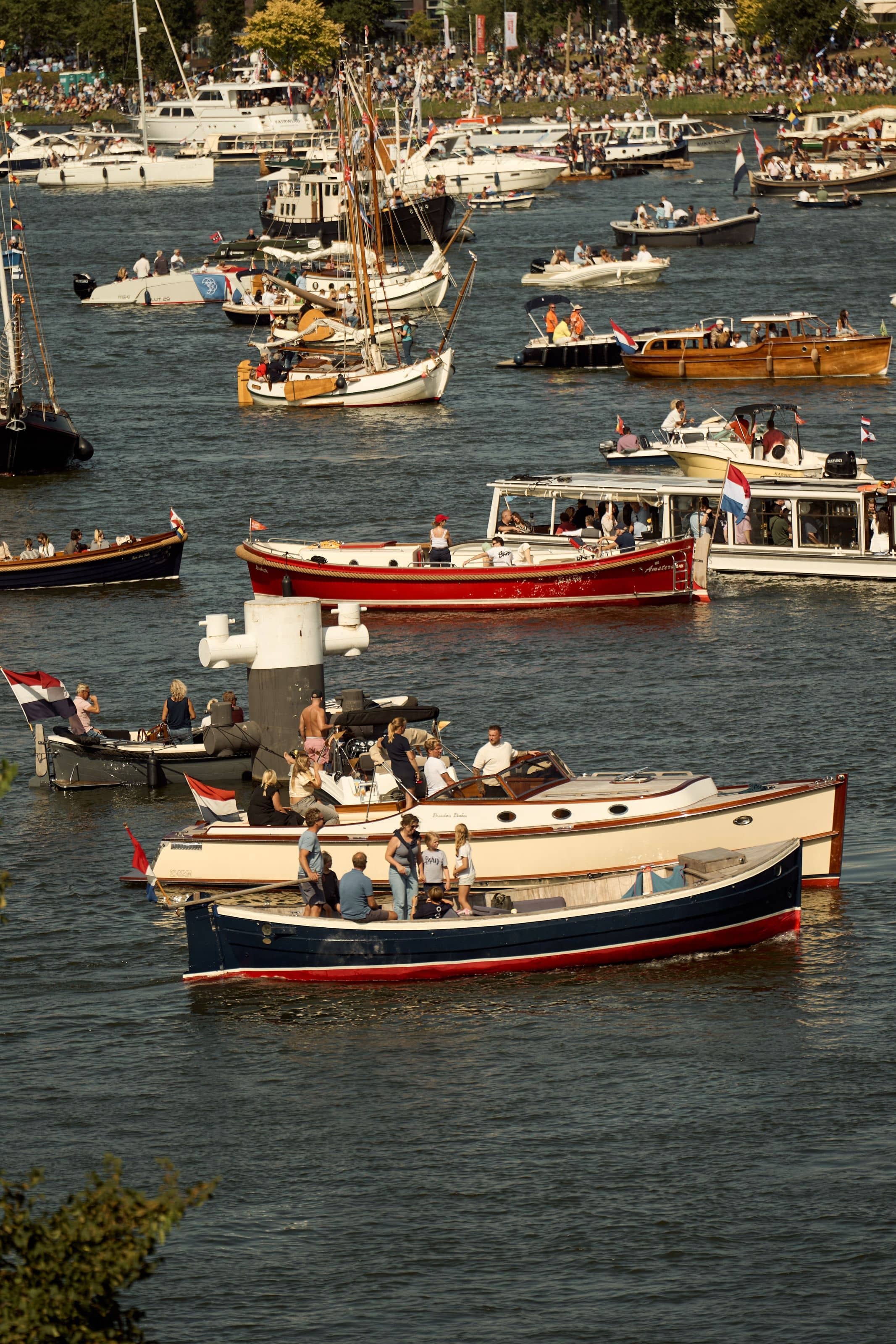 A dense flotilla of sloops, saloon boats and tenders crossing the IJ, people standing aboard in the afternoon light