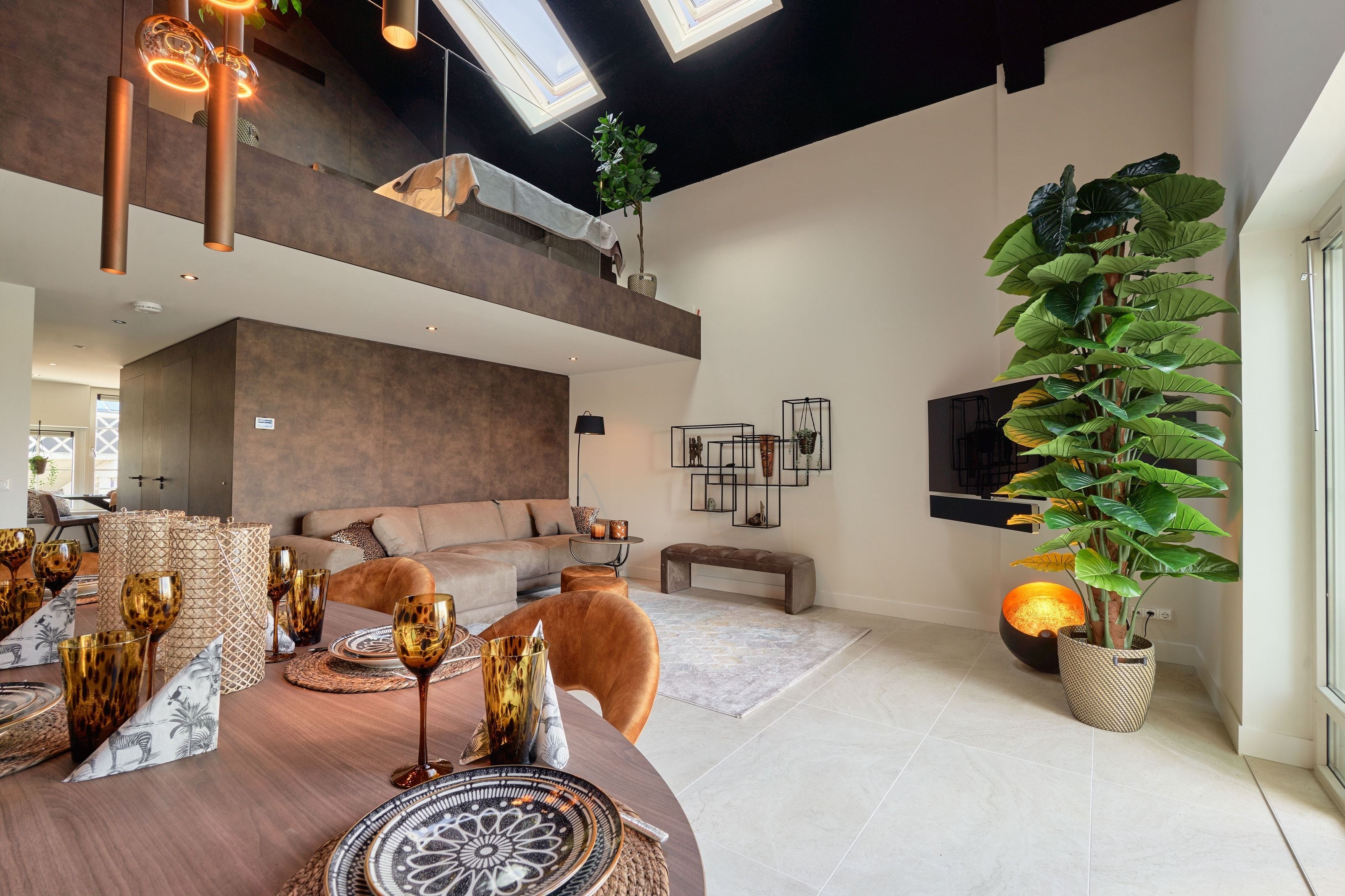 The double-height living space of the Nomad Water Lodge, a mezzanine bedroom behind glass under a black pitched ceiling, copper pendants over the dining table