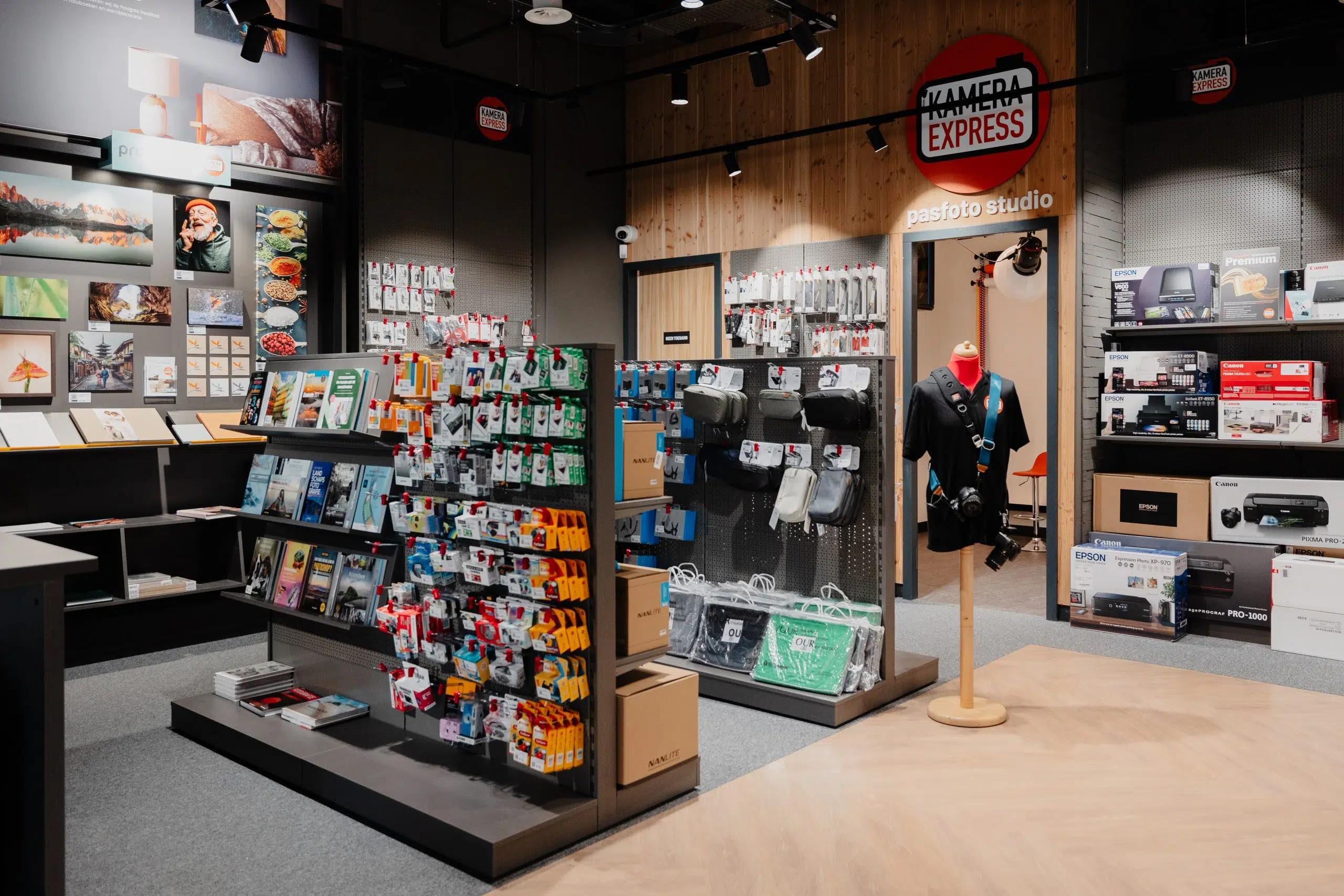 Inside a Kamera Express store: racks of ink and photo books, camera bags on pegboard, printer boxes along the wall and the passport-photo studio under the round logo
