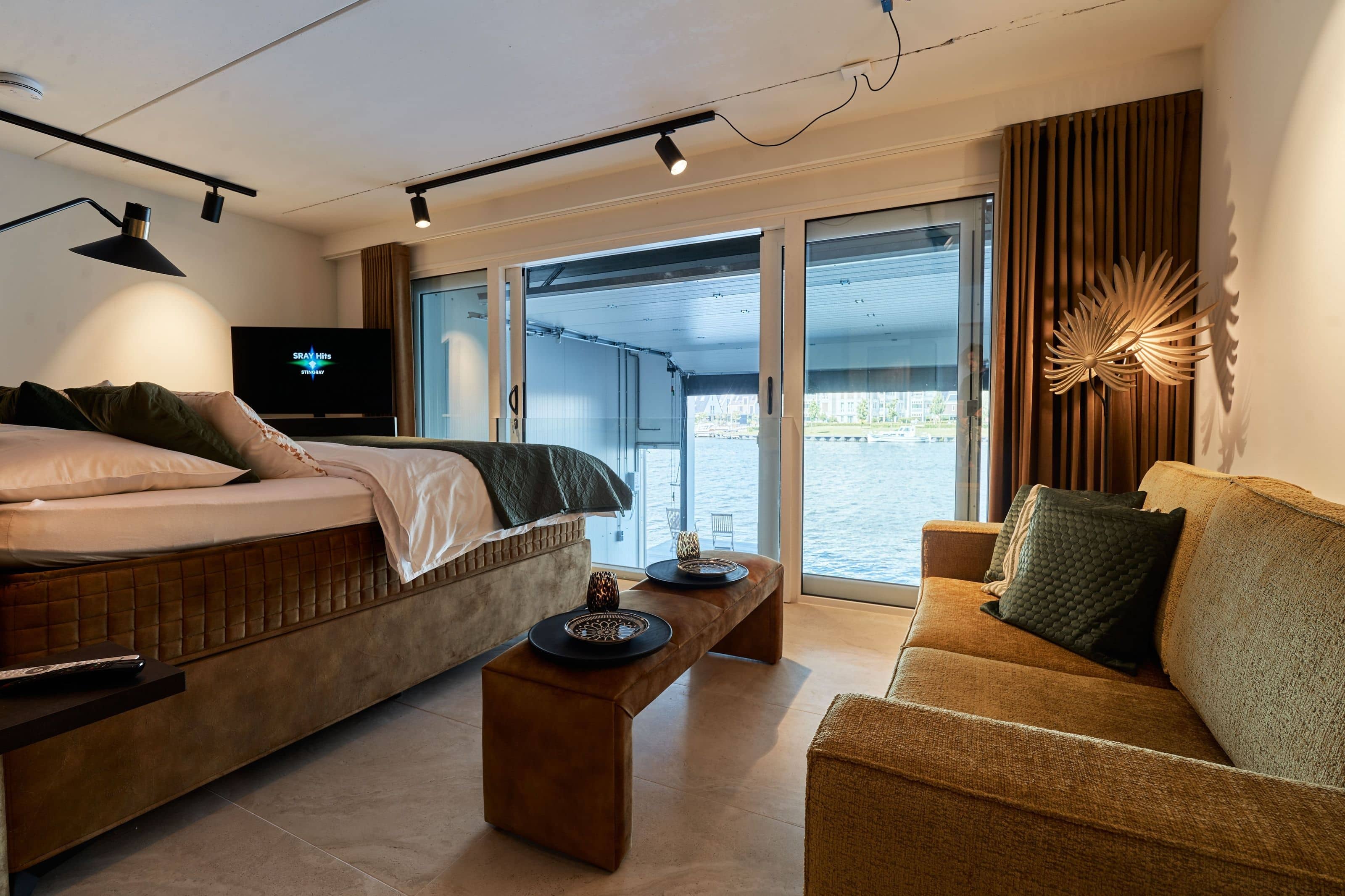 The Nomad Water Studio: a bed and a mustard sofa facing sliding doors that open straight onto the harbour