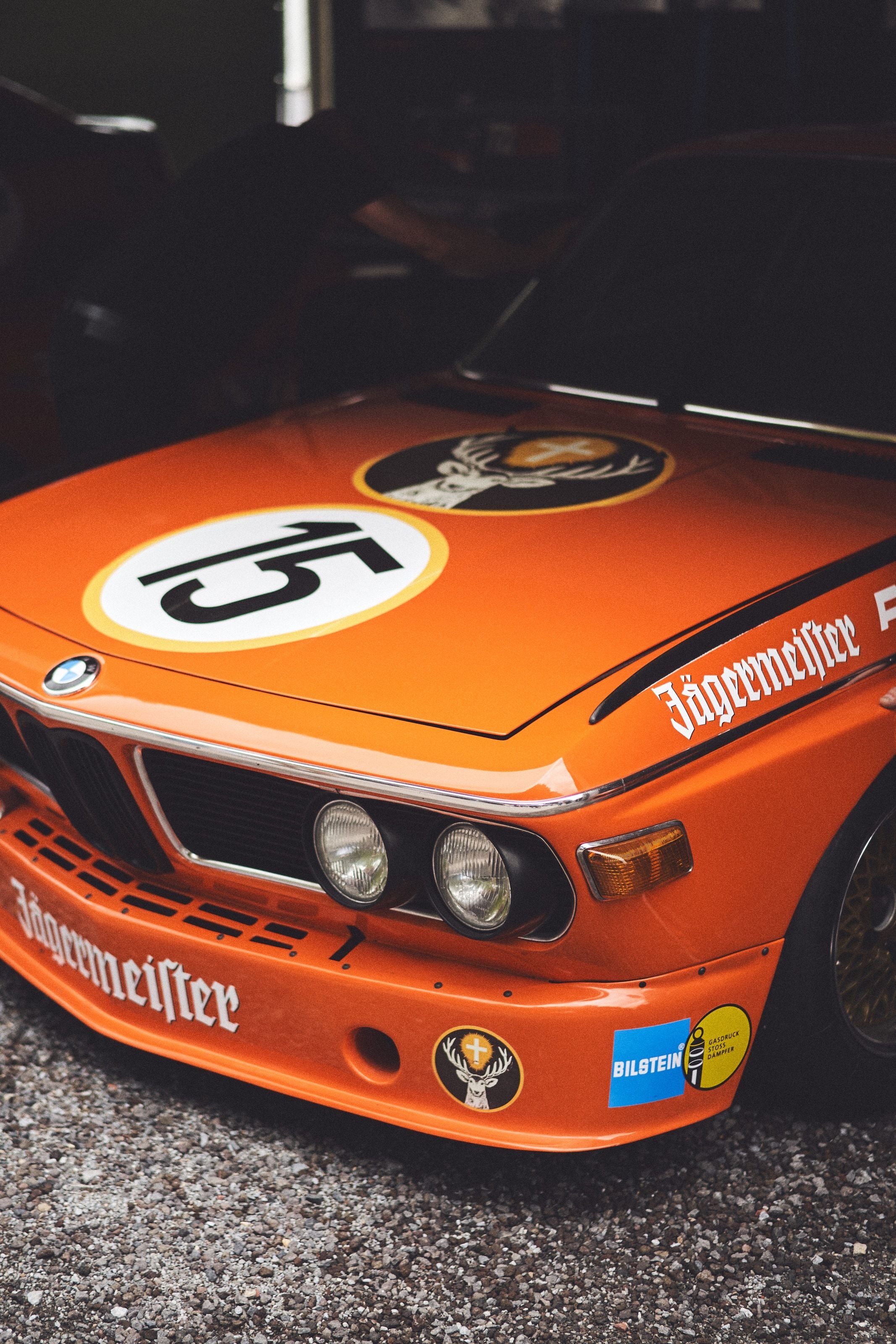 The orange Jägermeister-liveried nose of a BMW 3.0 CSL race car, number 15, lit dramatically in a dim garage