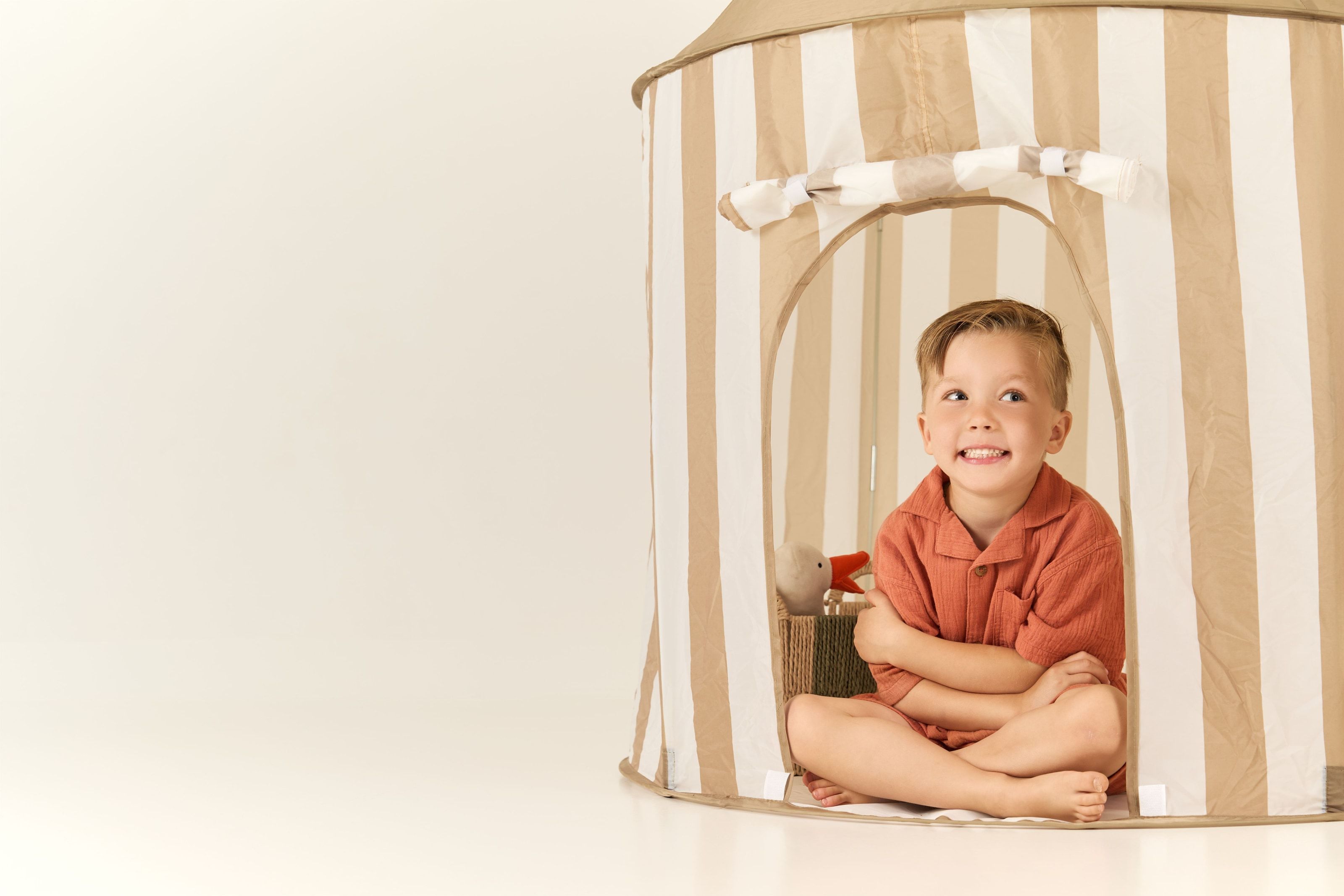 A boy sitting cross-legged in a striped play tent, grinning up out of the doorway, a toy goose in a basket beside him