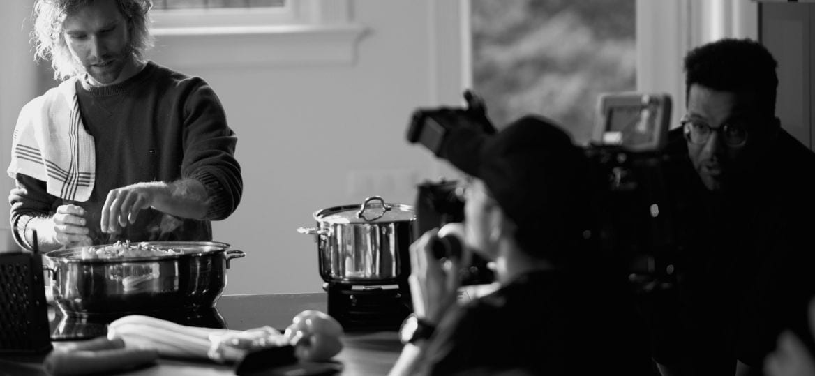 A cook working at a pan while a camera operator and a second crew member film him