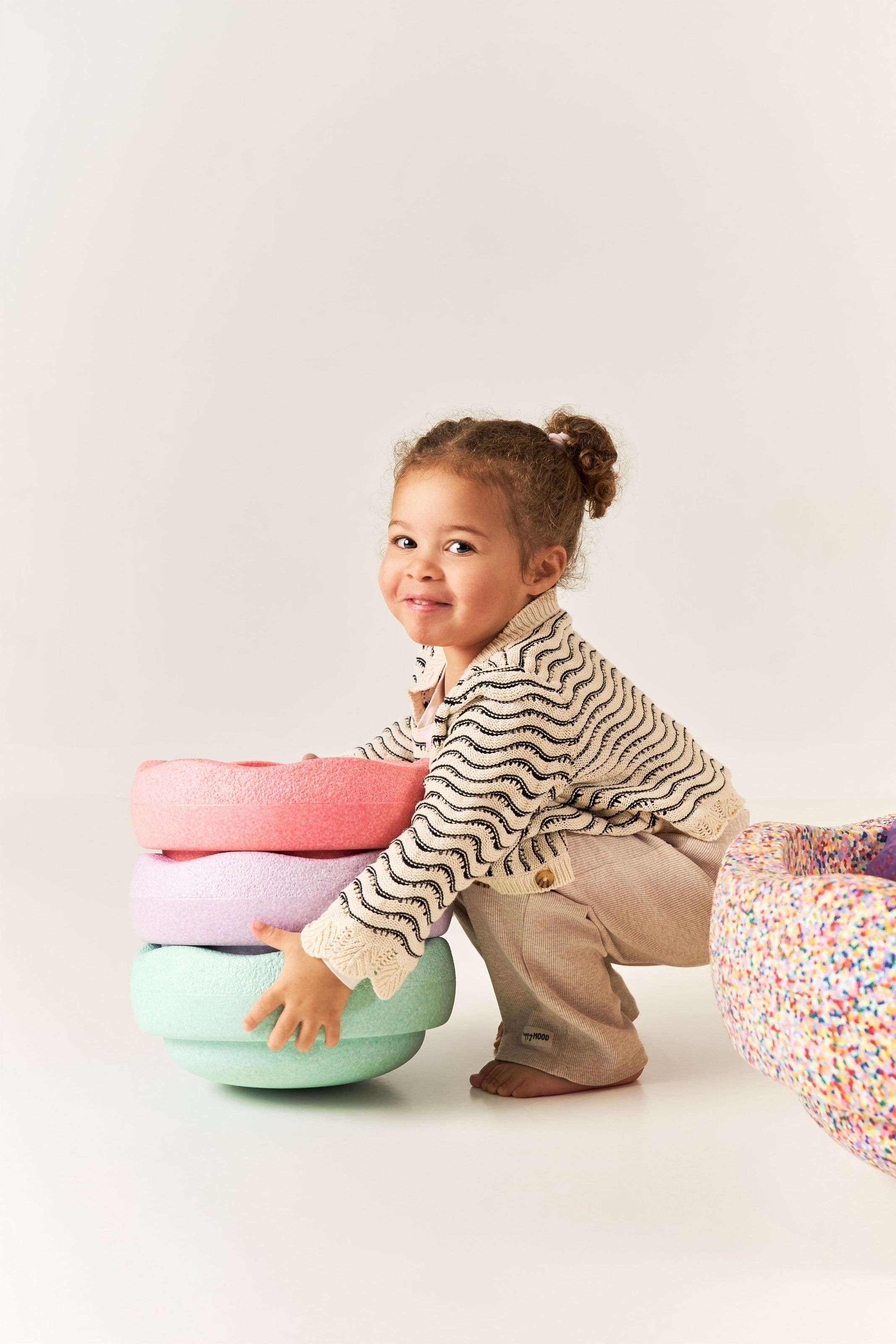 A toddler crouching to hug a stack of three pastel foam rings, looking round at the camera