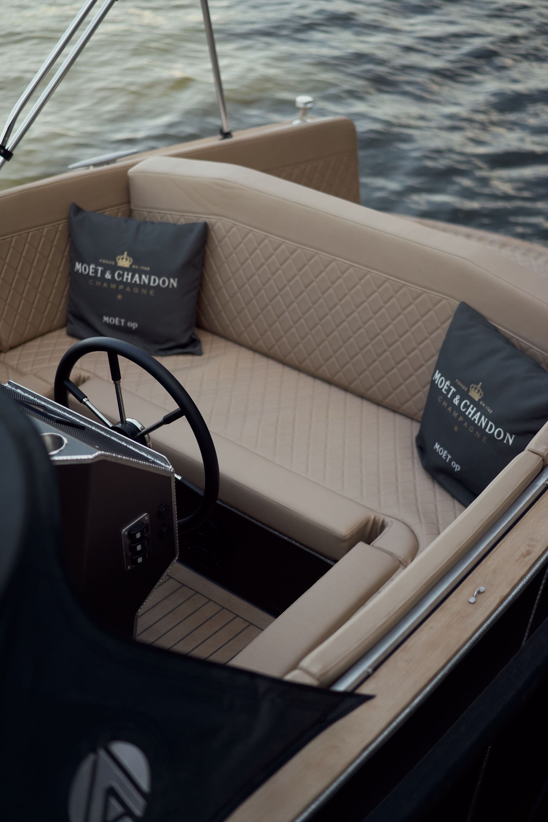 The tan leather seating and teak deck of the electric boat from above, two dark cushions printed Moët & Chandon set against the water