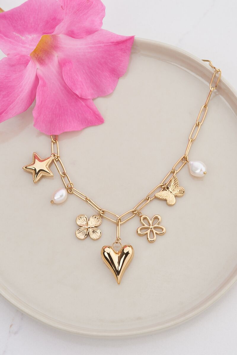 A gold charm necklace - a heart, a star, two flowers and a butterfly charm - laid beside a pink flower on a marble dish