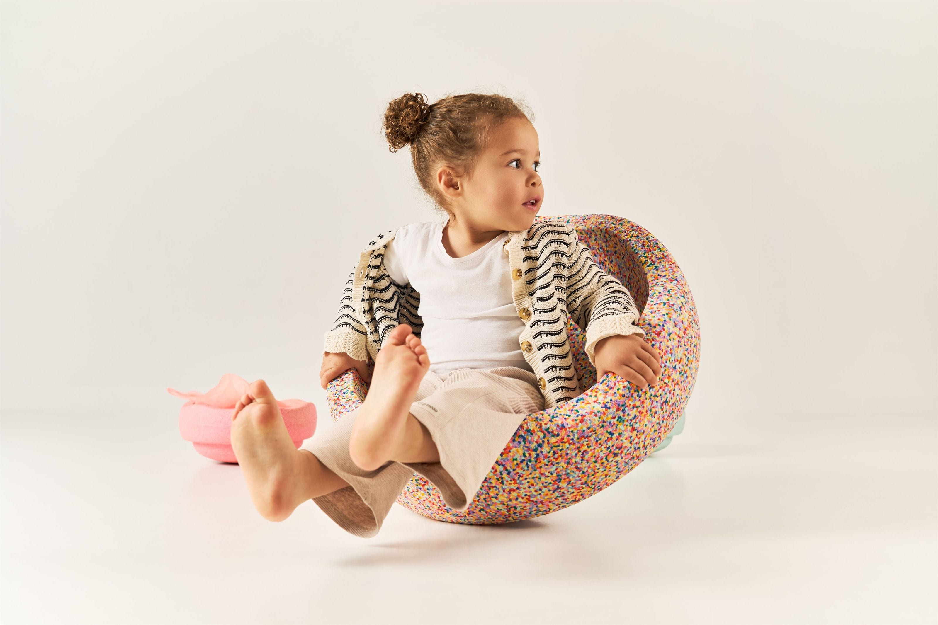 A toddler in a white t-shirt and striped cardigan sitting barefoot in a confetti-speckled foam rocker against a pale studio background