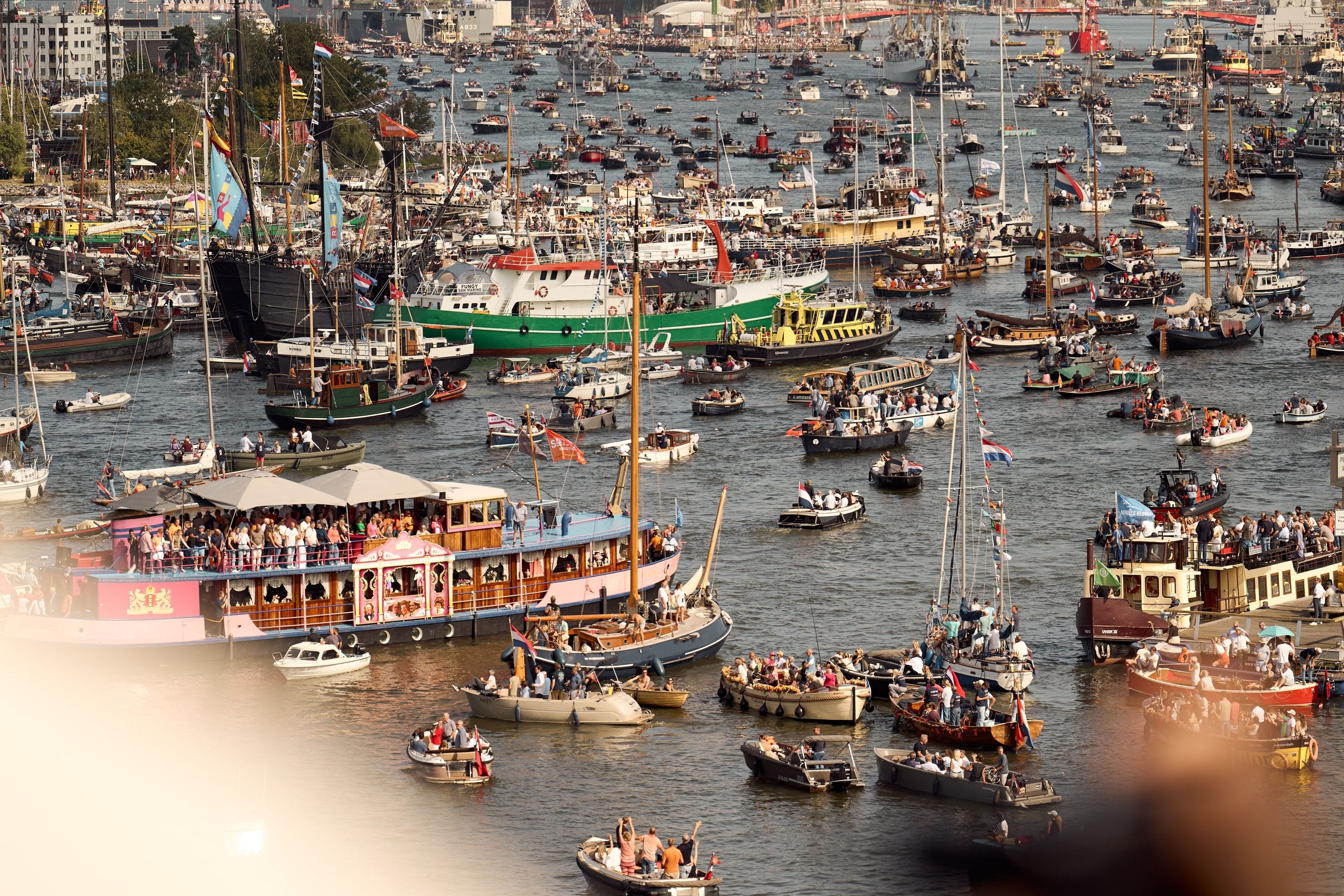 The IJ carpeted bank to bank with boats in late afternoon light, a pink and blue historic salon boat crowded with people in the foreground