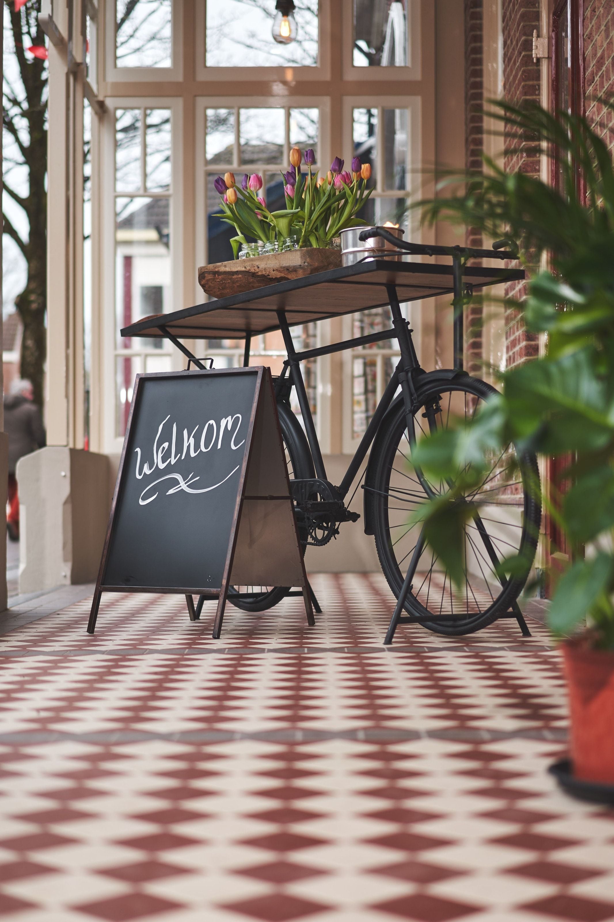 A chalked Welkom board beside a black delivery bike loaded with tulips, on the red and cream tiled entrance floor